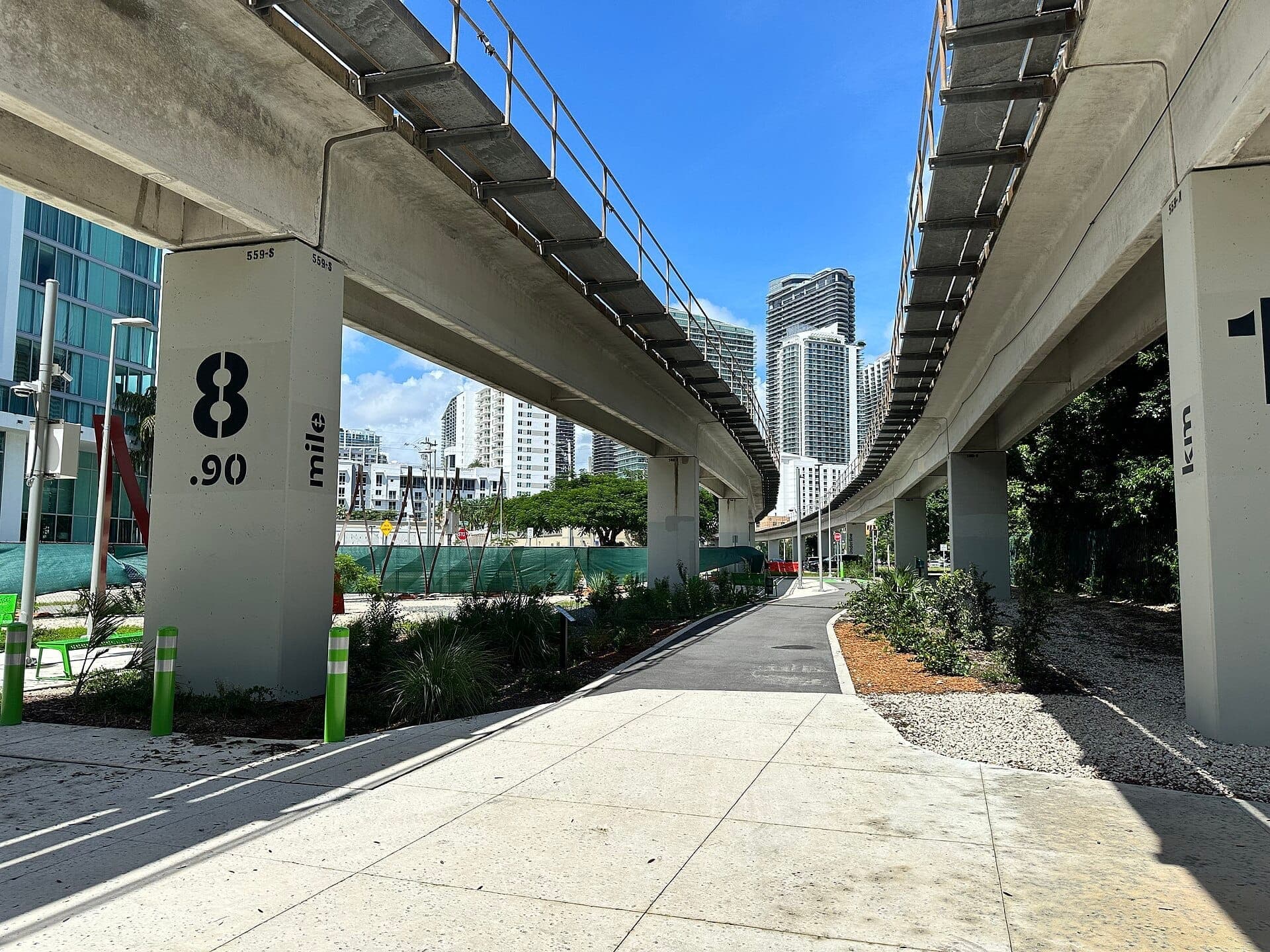 The Underline trail runs beneath Miami’s elevated Metrorail, surrounded by greenery, modern high-rise buildings, and clear blue skies.