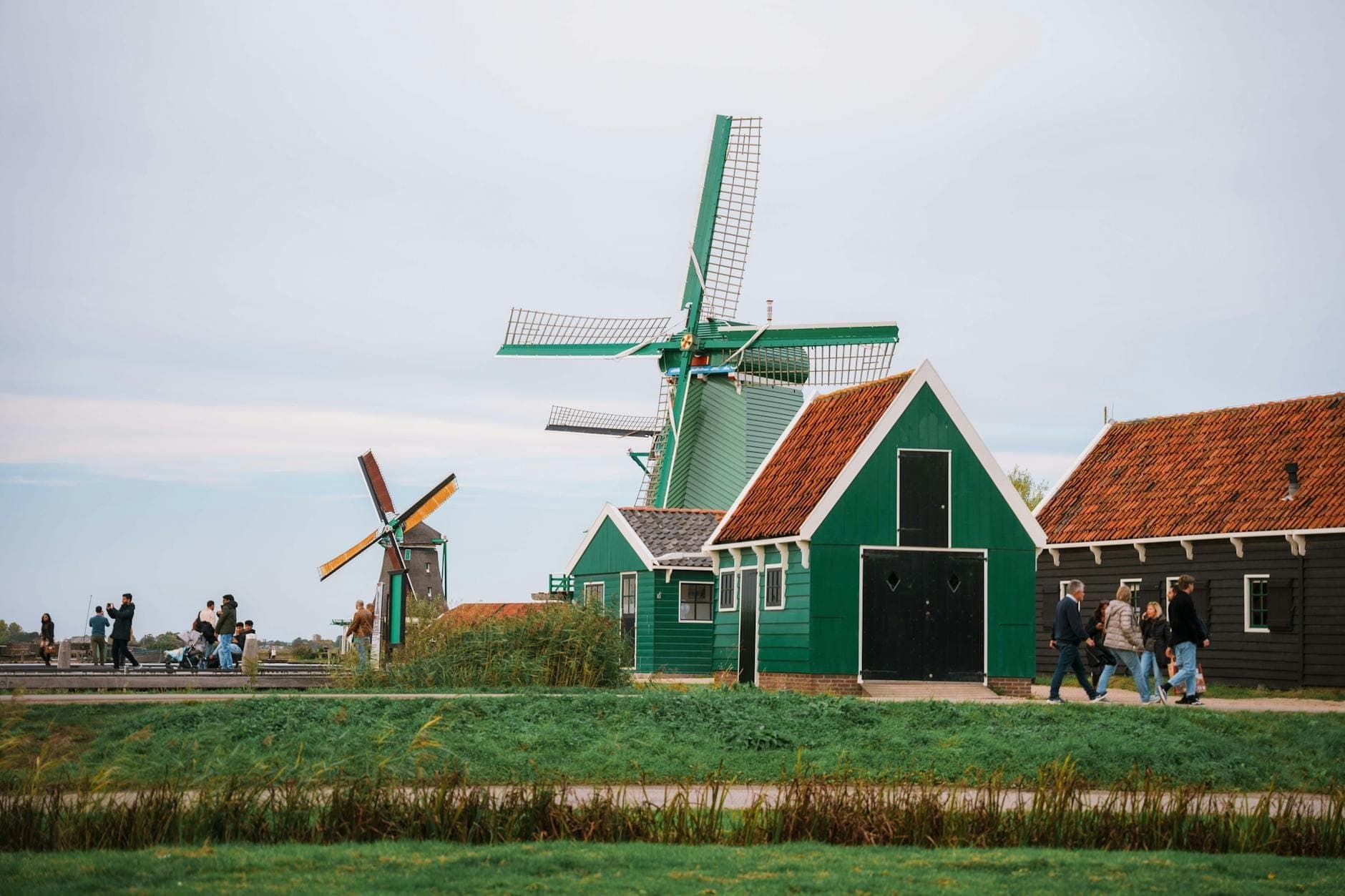 Touristen spazieren an traditionellen grünen Holzhäusern und funktionierenden Windmühlen am Wasser entlang, typisch für das Gebiet Zaanse Schans bei Amsterdam.