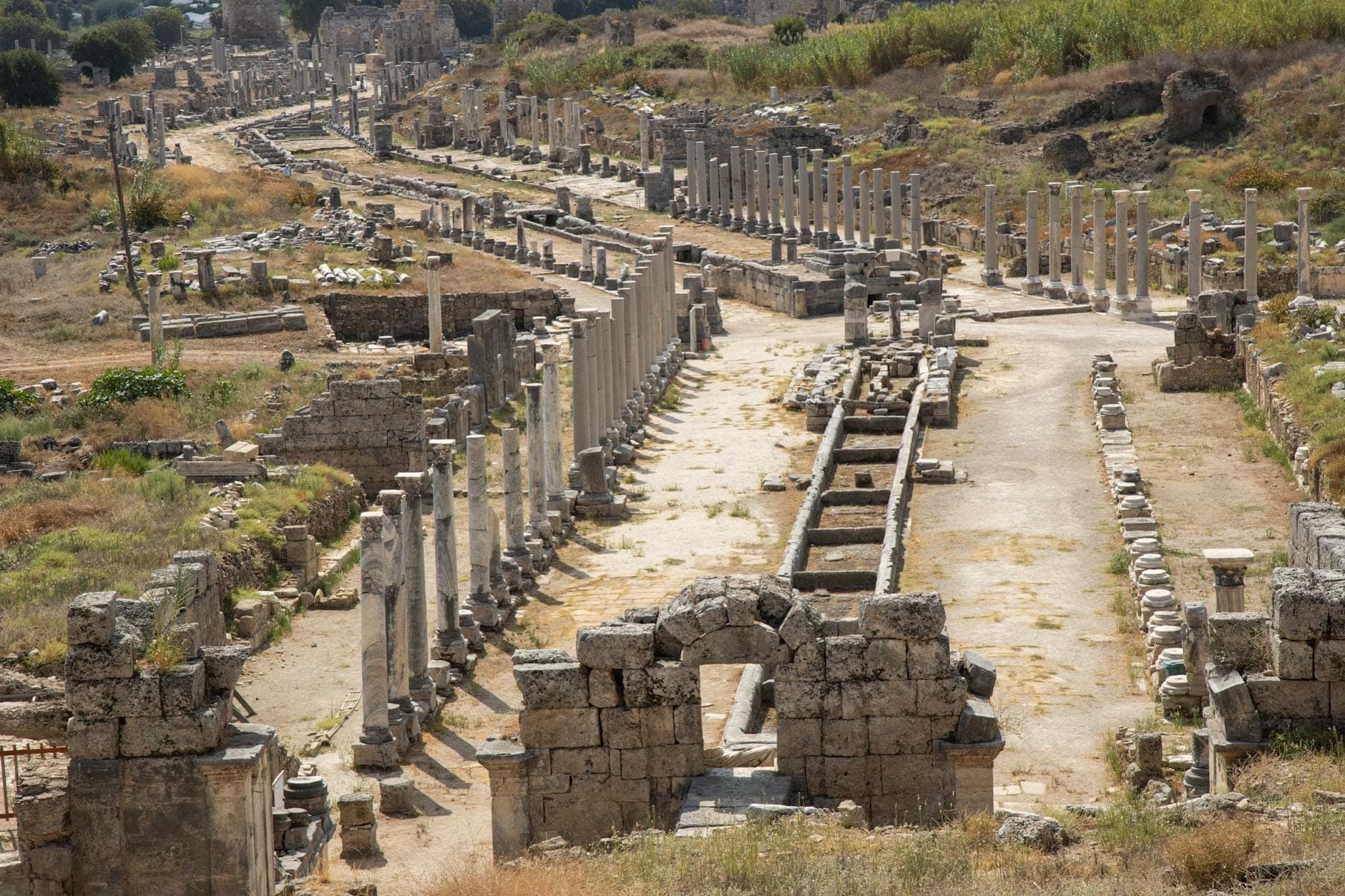 Vue panoramique des ruines d'une ville antique avec de longues rangées de colonnes en pierre et une maçonnerie éparpillée, sous un ciel dégagé près d'Antalya.