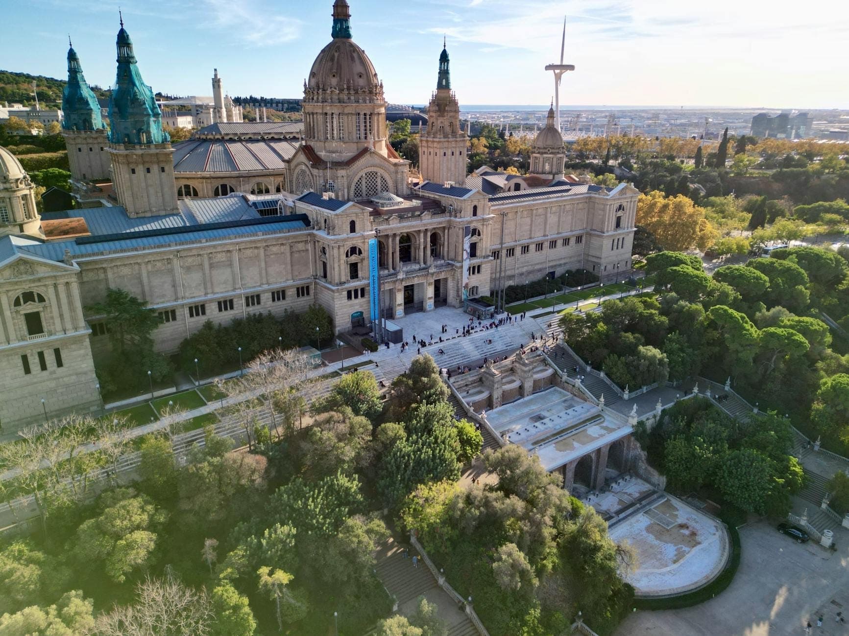 Vista aerea del Museo Nazionale d'Arte della Catalogna in cima alla collina di Montjuïc, circondato da giardini con la città sullo sfondo.