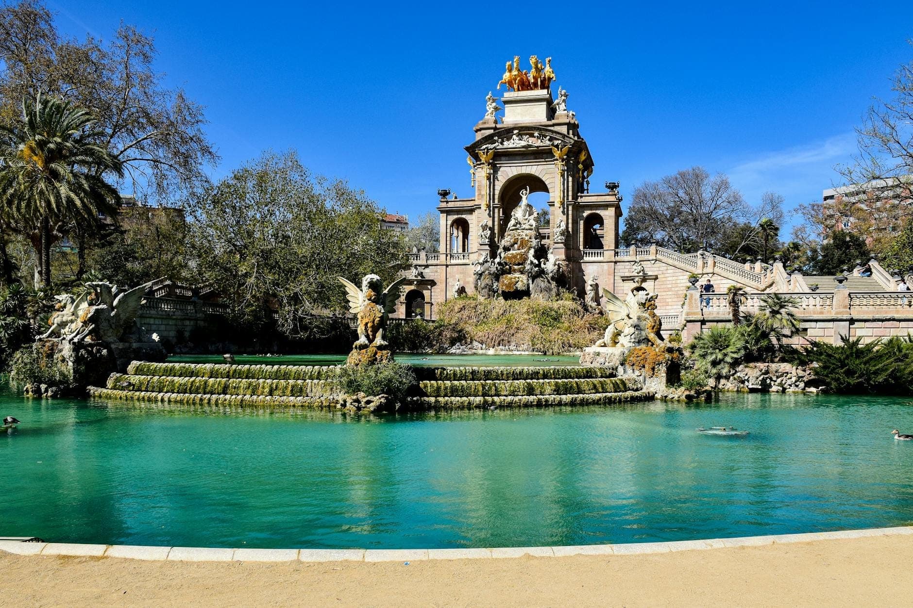 Vista ampia della fontana Cascada Monumental e del lago nel Parco della Ciutadella, Barcellona, sotto un cielo azzurro brillante.