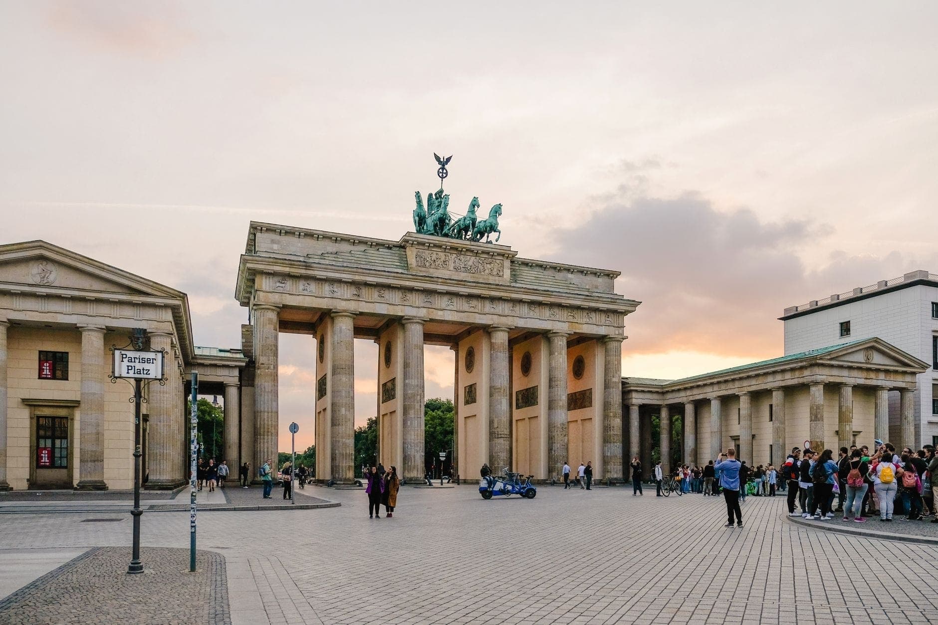 Vista nocturna de la Puerta de Brandeburgo en Berlín con turistas en la plaza y un colorido cielo al atardecer de fondo.
