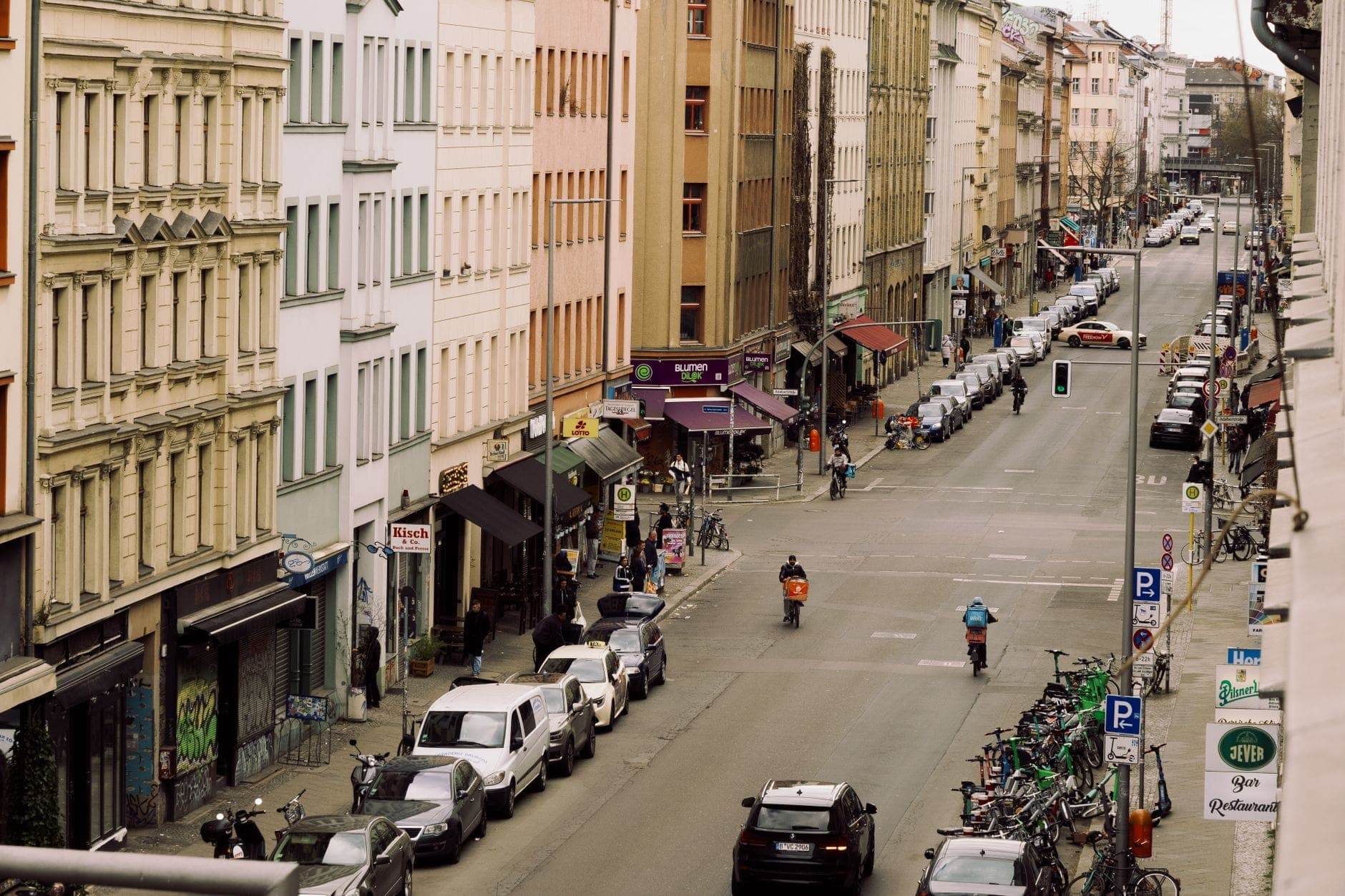Una animada escena callejera de Berlín vista desde arriba con coloridos edificios, tiendas, bicicletas, coches aparcados y personas caminando y en bicicleta.