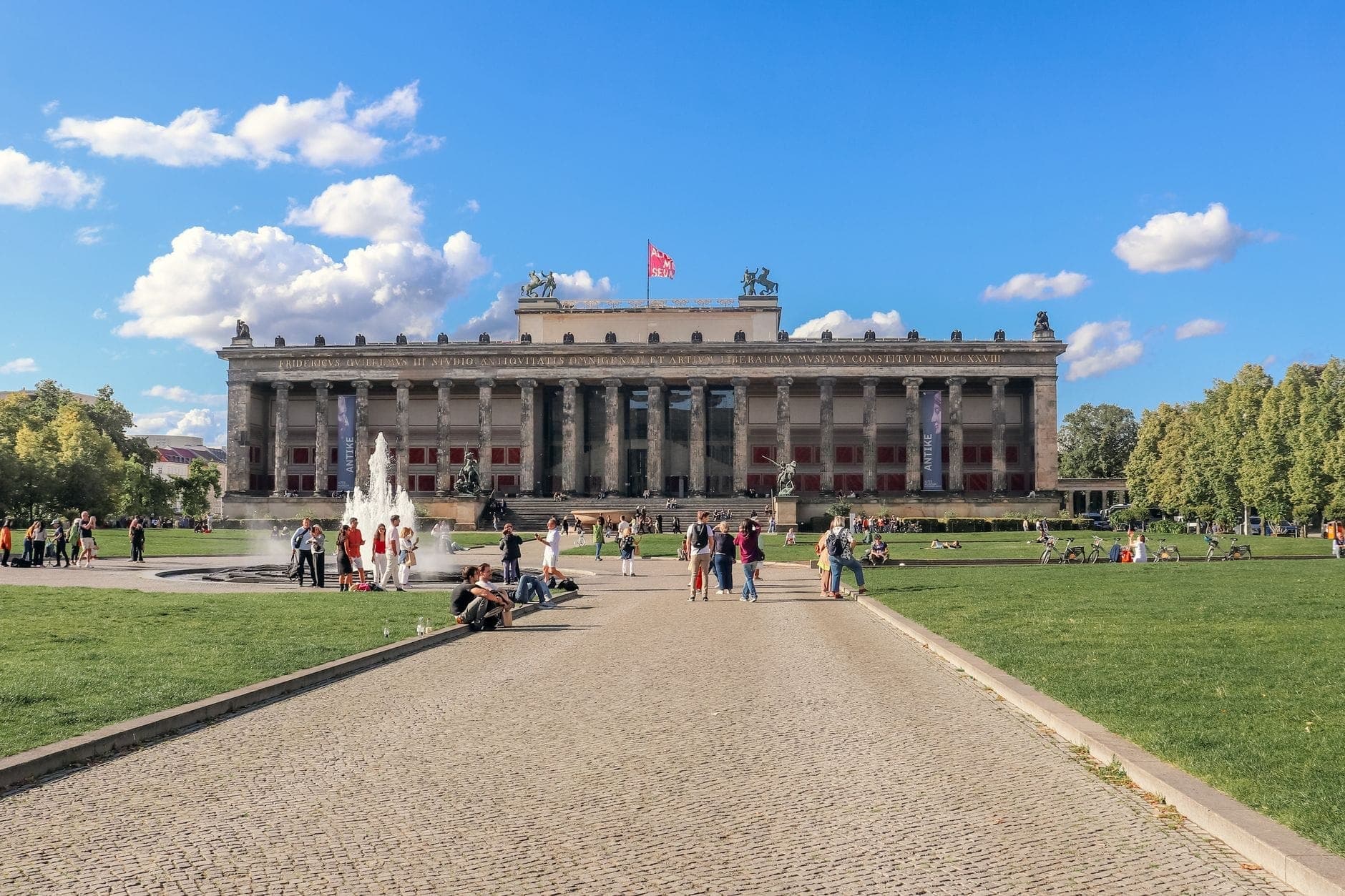 Vista amplia del Altes Museum en la Isla de los Museos de Berlín con personas descansando en céspedes verdes y una fuente al frente en un día soleado