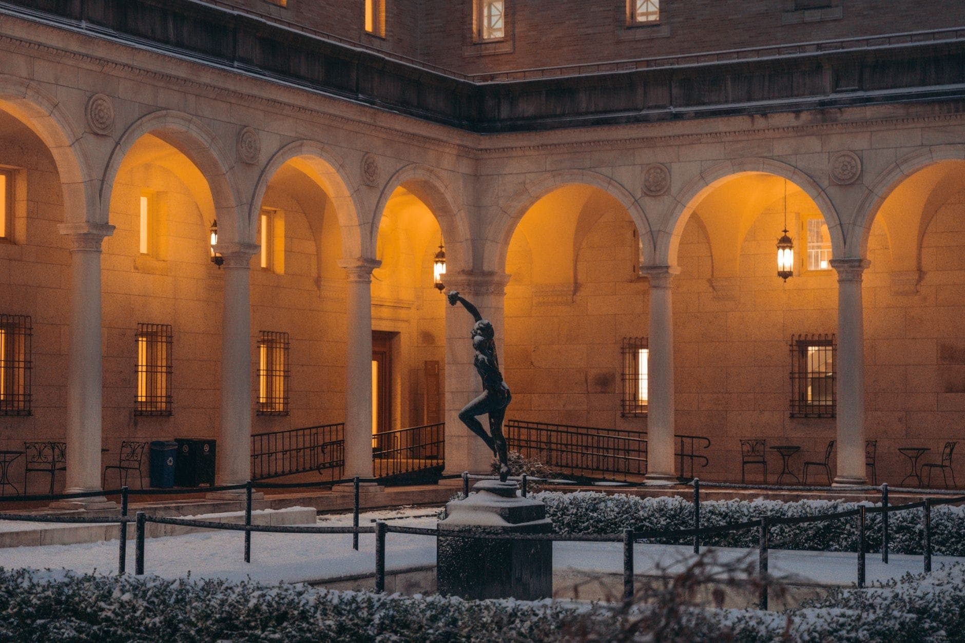 Cour d'un musée de Boston au crépuscule, avec des arches, des lanternes brillantes et une statue au centre saupoudrée de neige.