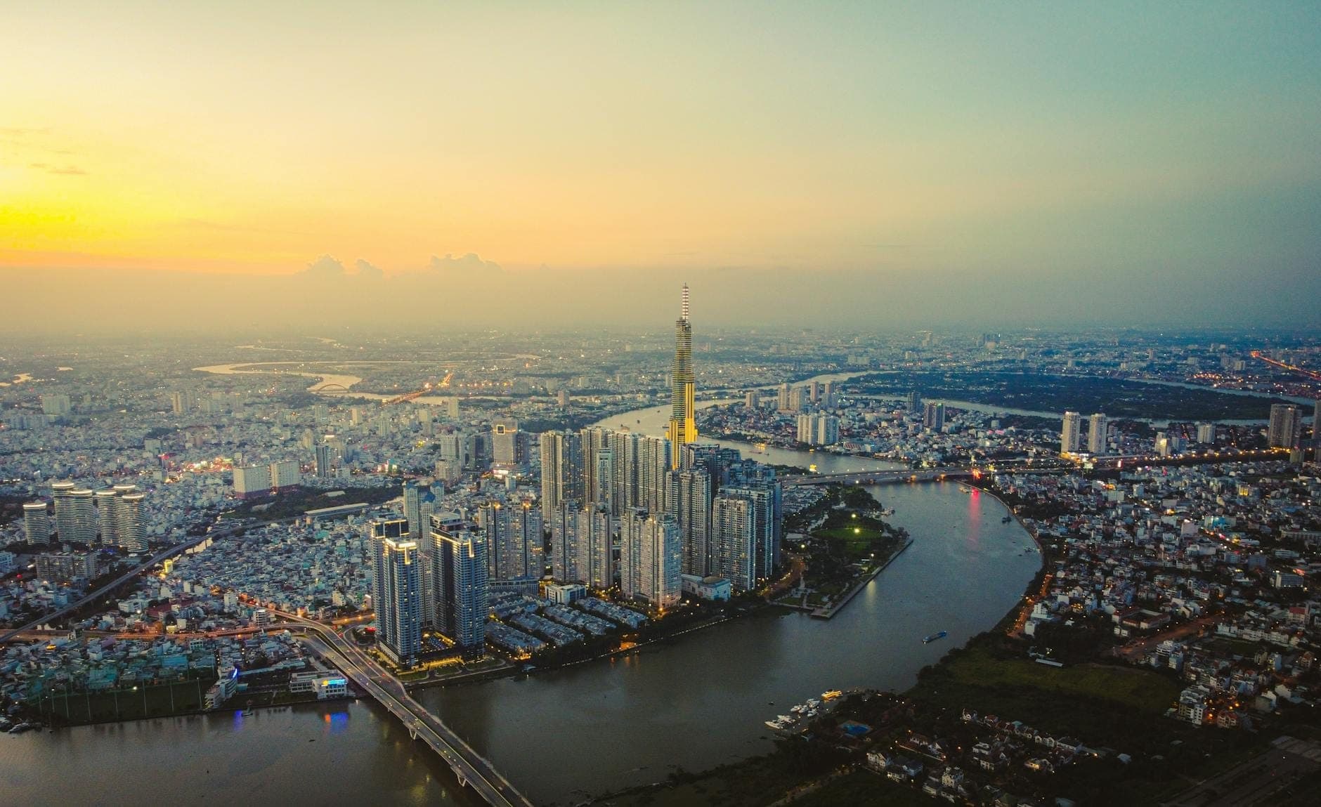 Aerial na tanawin ng skyline ng Ho Chi Minh City na may tore ng Landmark 81, kurbada ng ilog, at mga gusali sa paglubog ng araw sa ilalim ng makulay na langit.