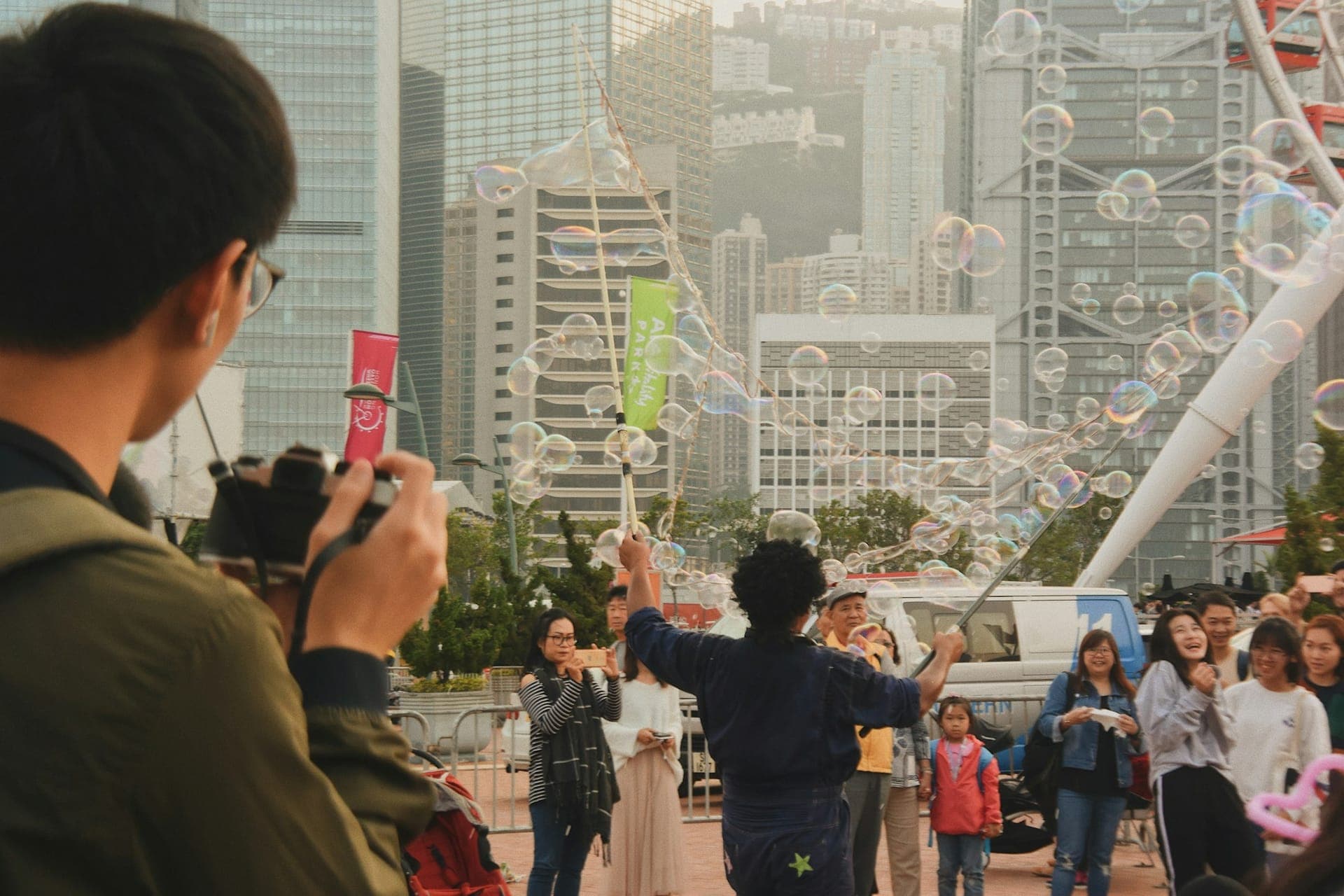 Street performer blowing giant soap bubbles to a crowd in Hong Kong's Central district with modern skyscrapers rising behind Victoria Harbour.
