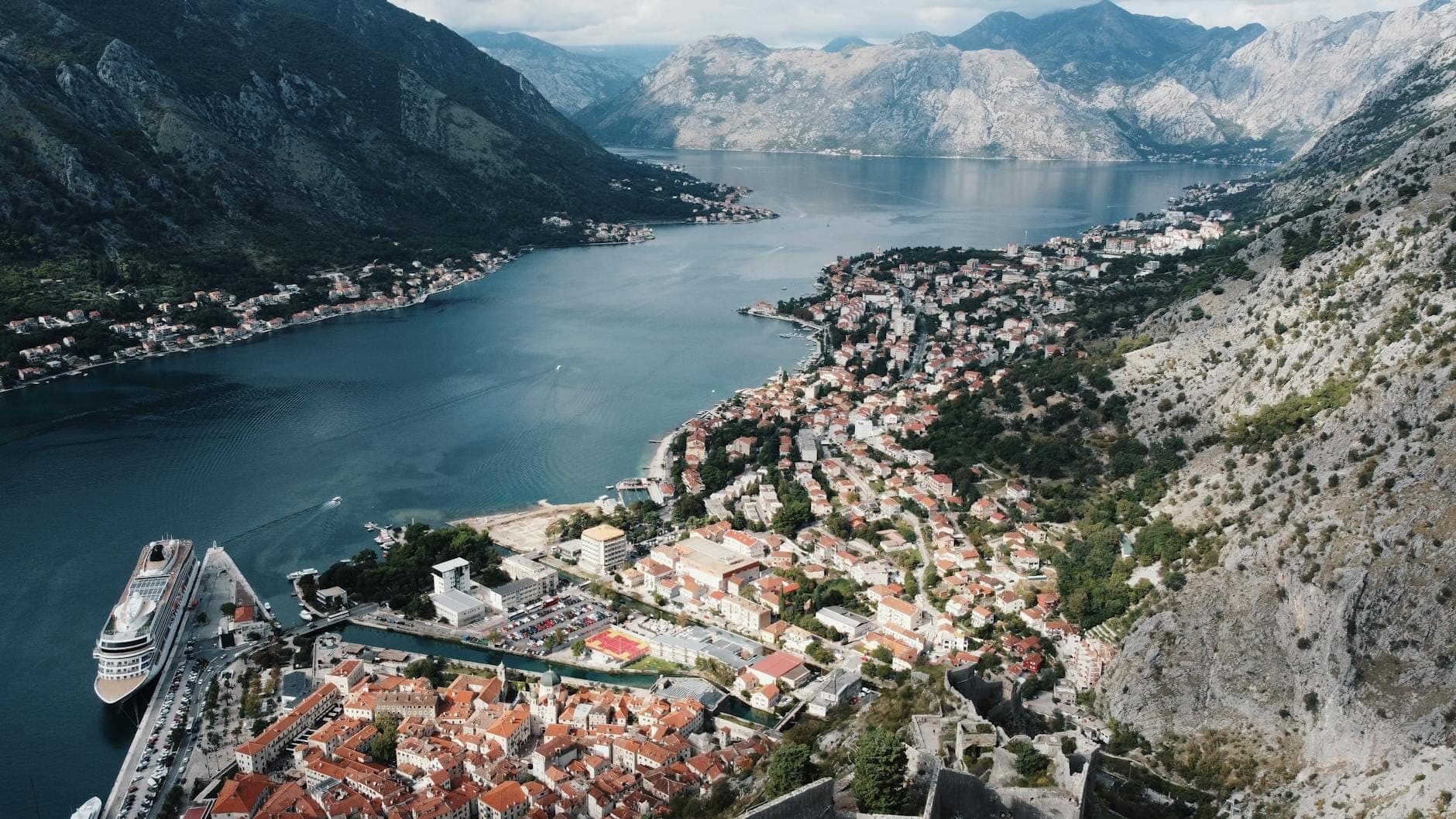 Vue aérienne de Kotor avec la vieille ville, un paquebot de croisière, le front de mer et les montagnes spectaculaires encerclant la baie sous un ciel partiellement nuageux.