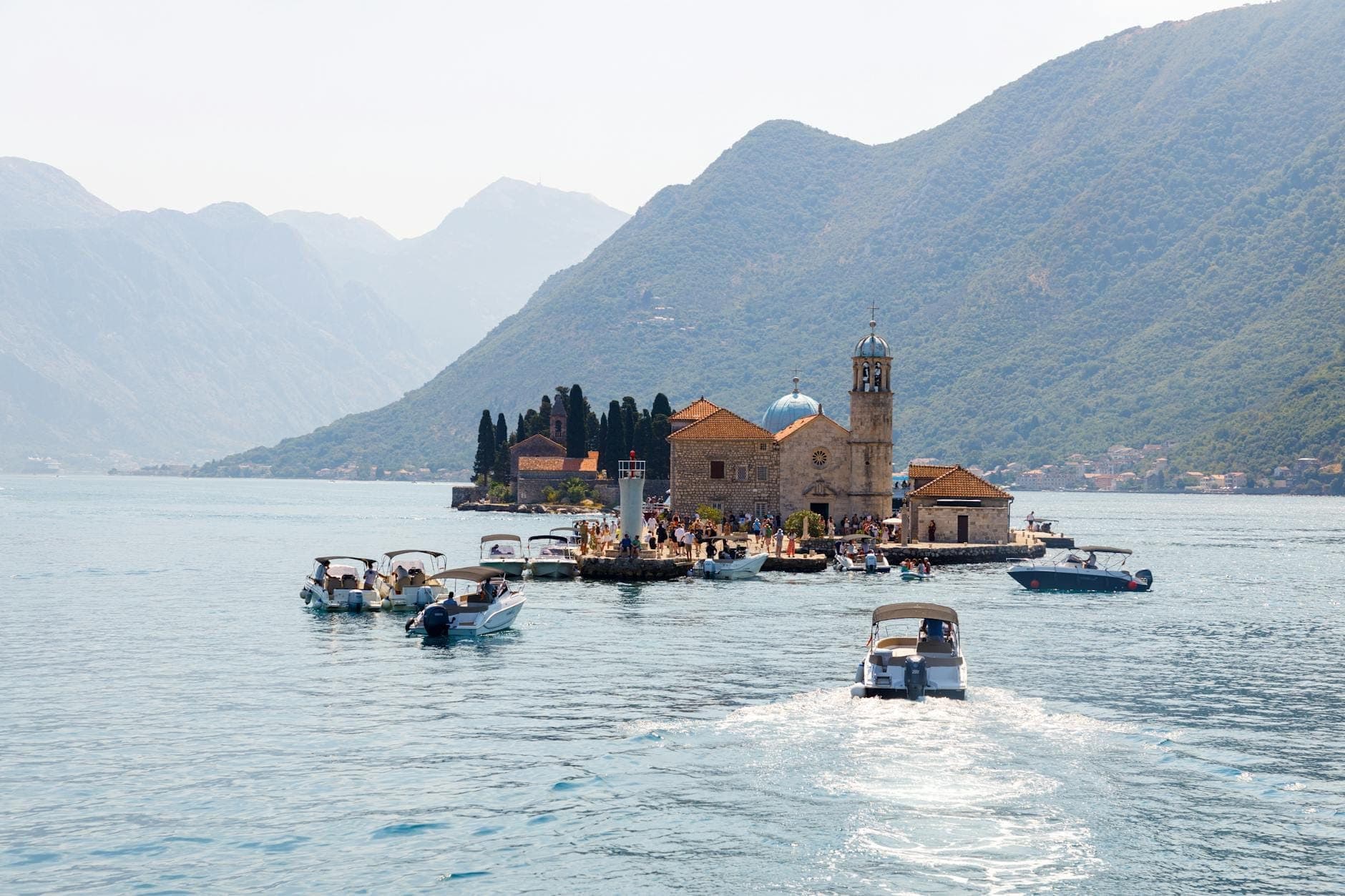 Plusieurs bateaux approchent la petite île de Our Lady of the Rocks dans la baie de Kotor, entourée de montagnes et d'une eau bleue et claire.