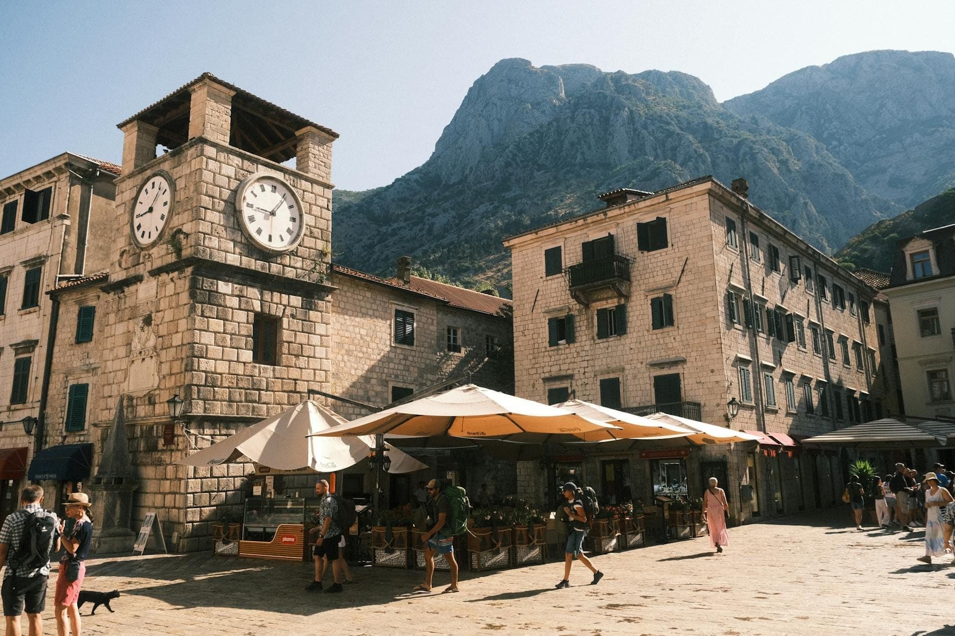 Grande place dans la vieille ville de Kotor avec la célèbre tour de l'horloge en pierre, des bâtiments historiques, des cafés en plein air et des personnes se promenant sous des parasols, avec des montagnes en arrière-plan.