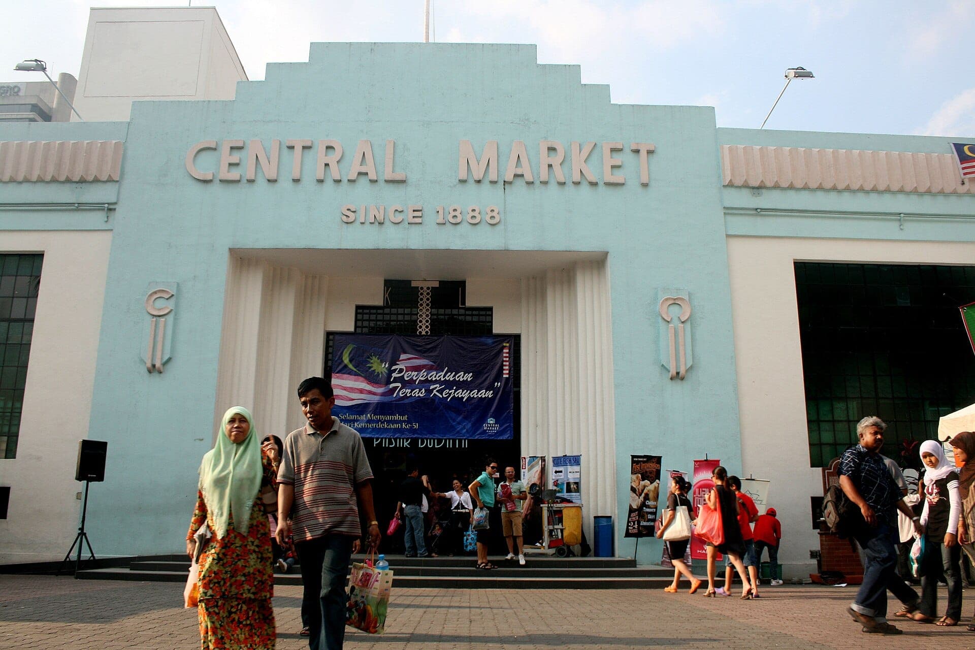 Central Market в Куала-Лумпуре с характерным синим фасадом в стиле ар-деко, прохожими и ярким послеполуденным солнечным светом.