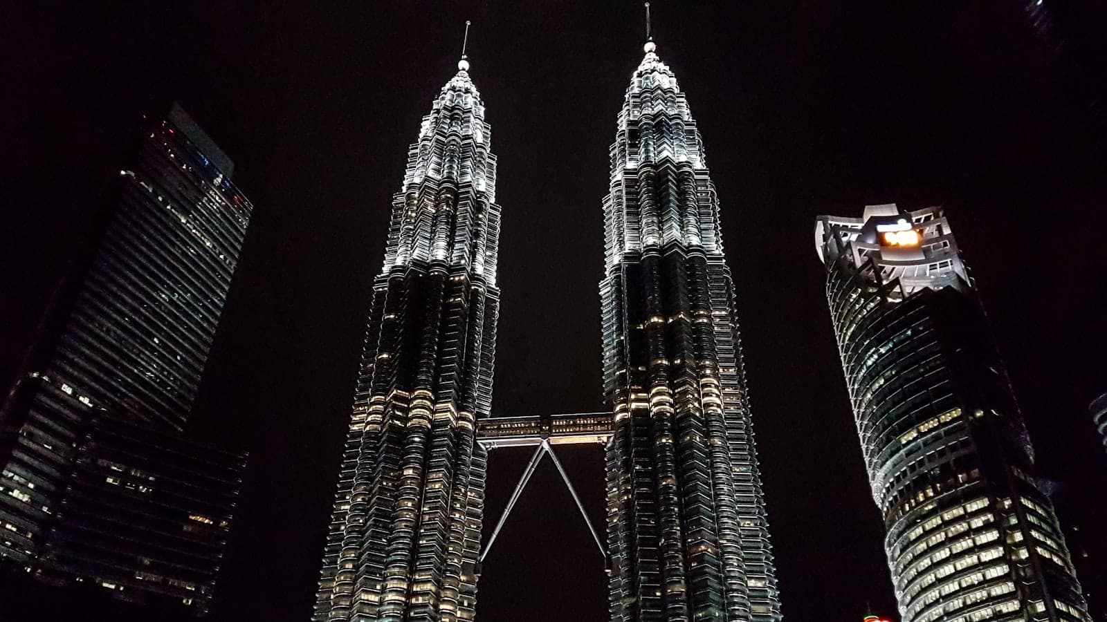 Petronas Twin Towers в Куала-Лумпуре, освещённые ночью, в окружении других современных небоскрёбов на фоне тёмного неба.