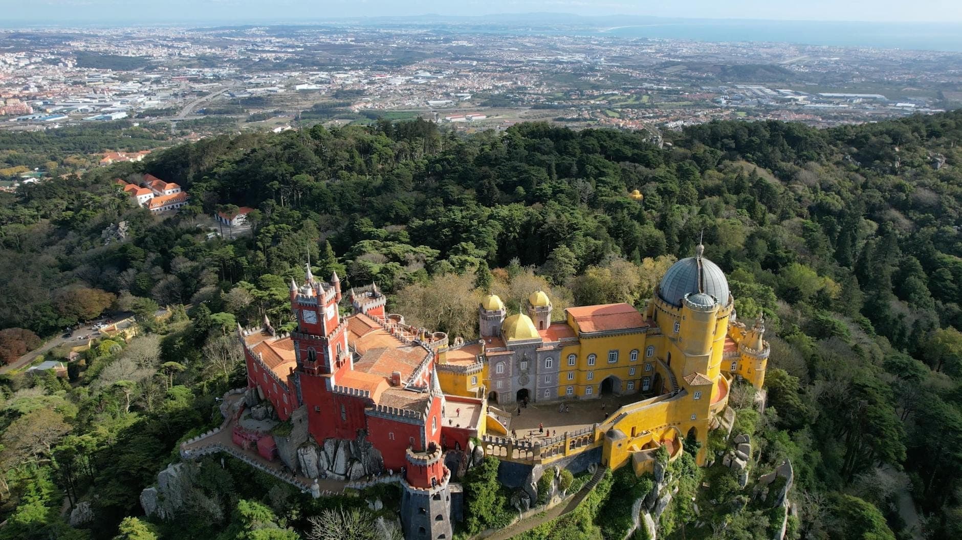 Vista aérea do Palácio da Pena rodeado por colinas arborizadas exuberantes com Lisboa e o Atlântico ao fundo.