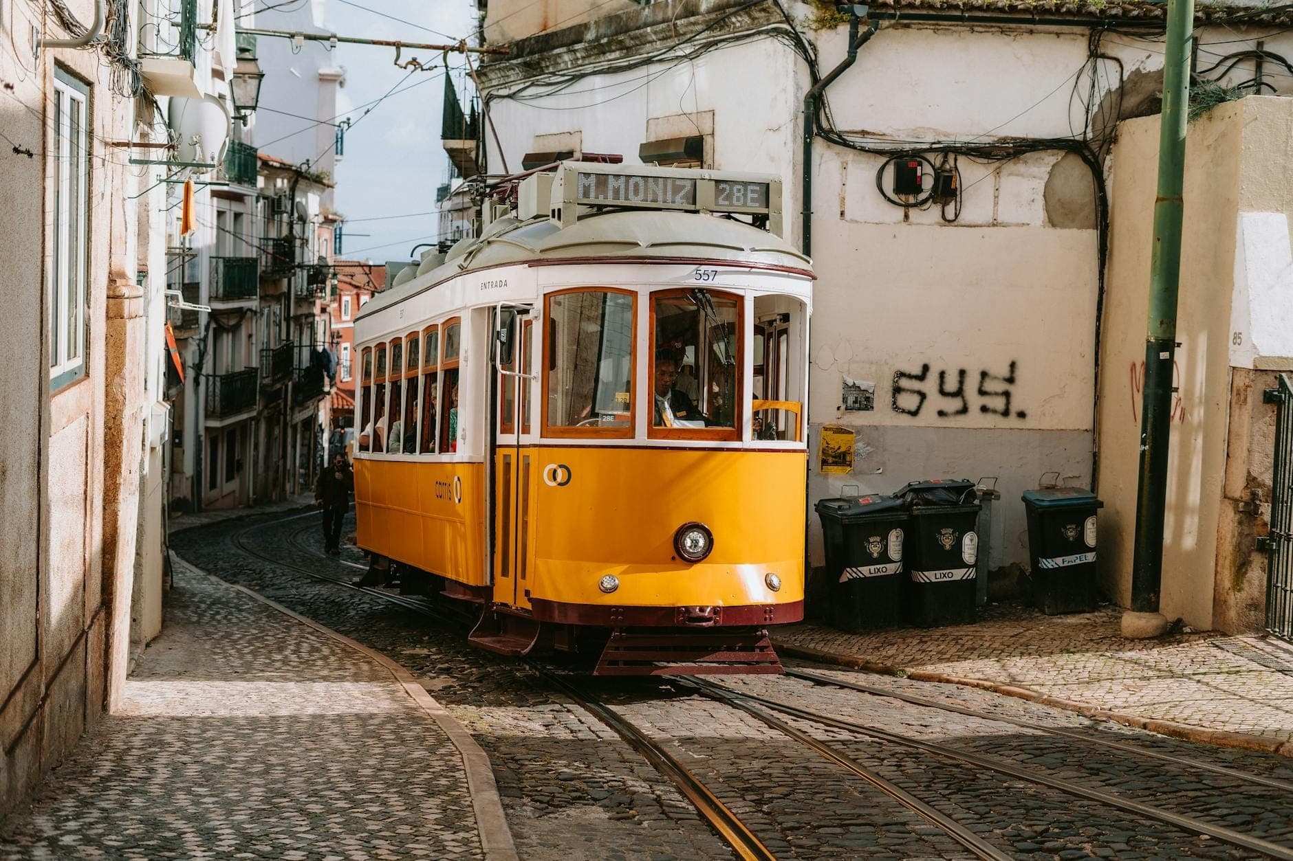 Um clássico elétrico amarelo de Lisboa percorre uma rua estreita, íngreme e de calçada portuguesa, ladeada por edifícios antigos em Alfama ou Graça.