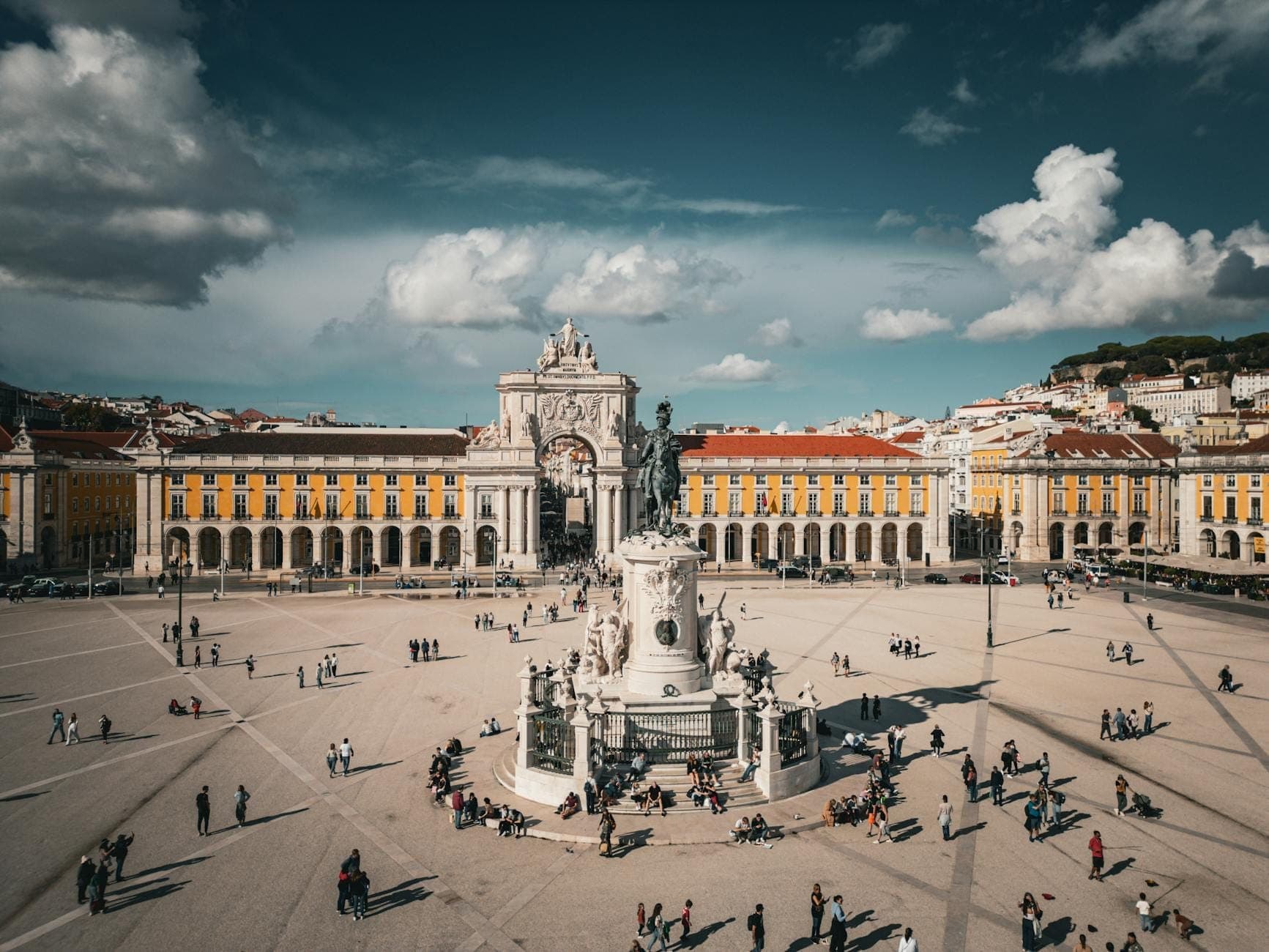 Praça do Comércio no centro de Lisboa com o Arco da Rua Augusta, uma estátua e pessoas caminhando pela grande praça em um dia ensolarado.