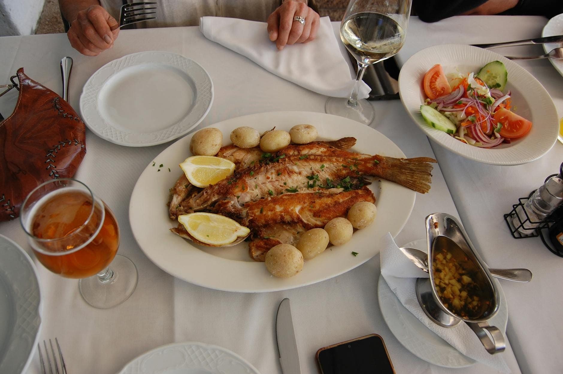 Uma mesa posta com pratos de peixe grelhado, batatas, salada, vinho e cerveja; as mãos dos comensais são visíveis, sugerindo uma refeição num restaurante português.