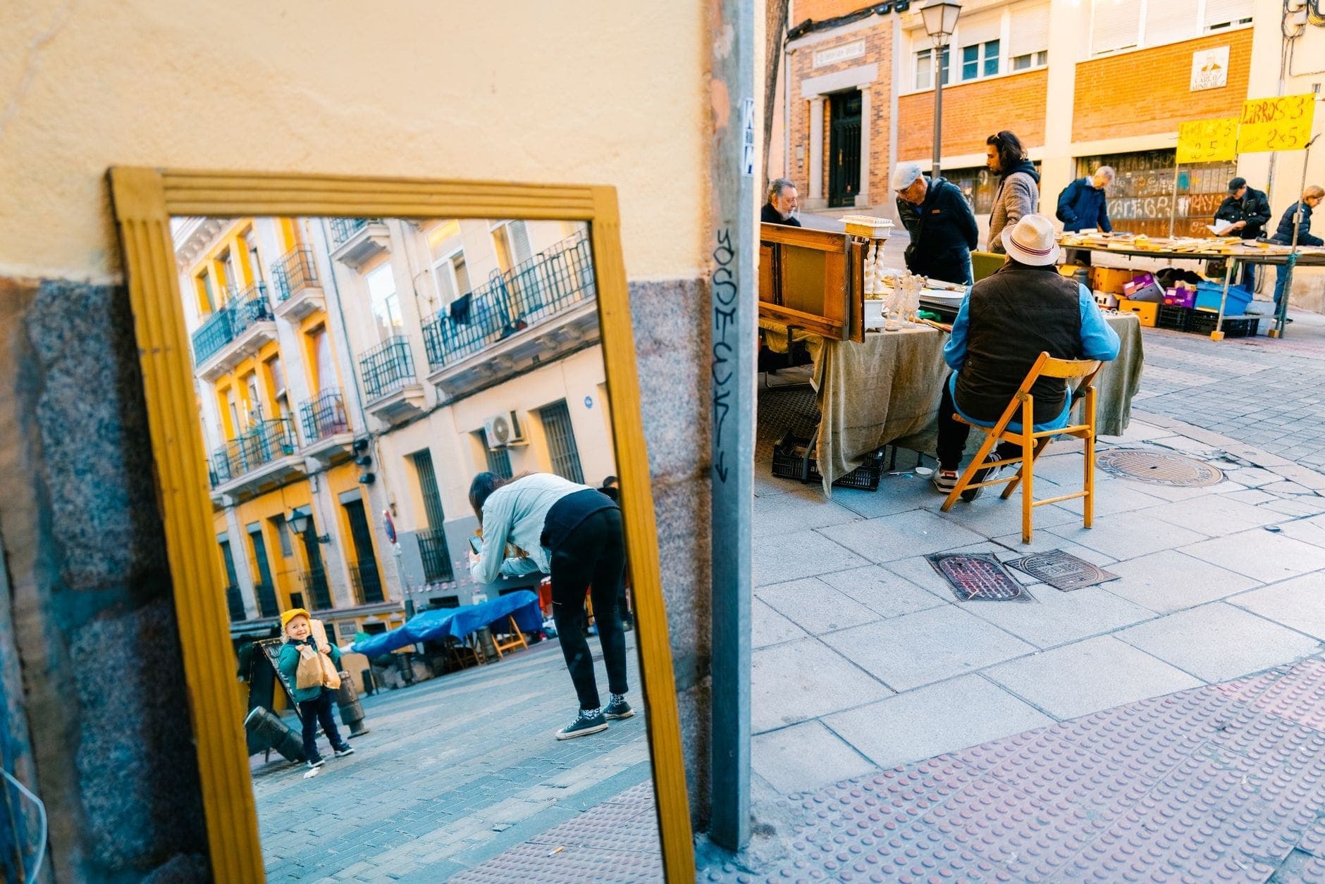 Buntes Straßenmarktgeschehen in Madrid mit Menschen, die Stände durchstöbern, einem Spiegel, der das Stadtleben widerspiegelt, und historischen Gebäuden des Viertels.