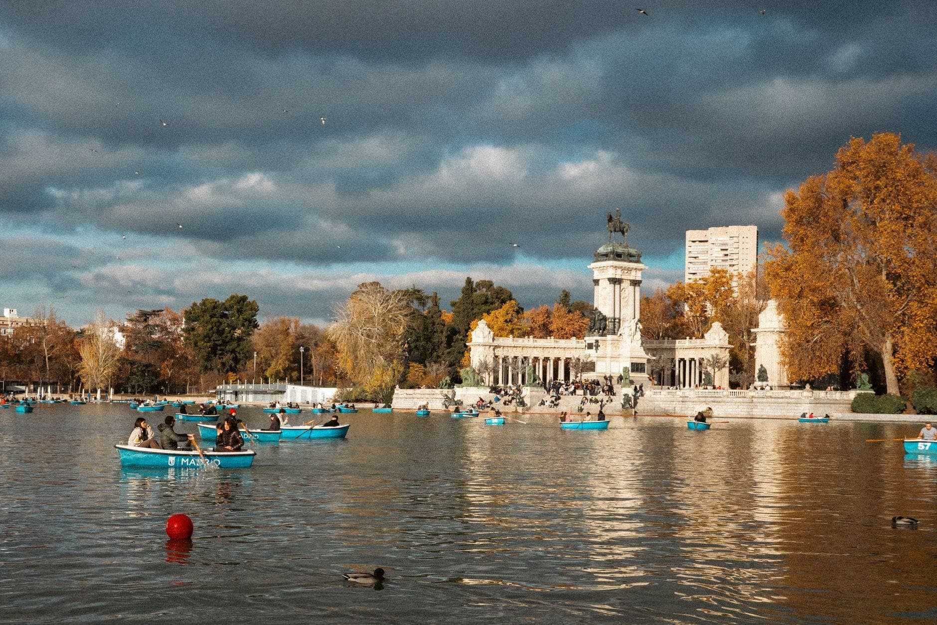 Menschen rudern in blauen Booten auf dem großen Teich vor dem Monumento a Alfonso XII im Parque del Retiro in Madrid an einem bewölkten Tag.