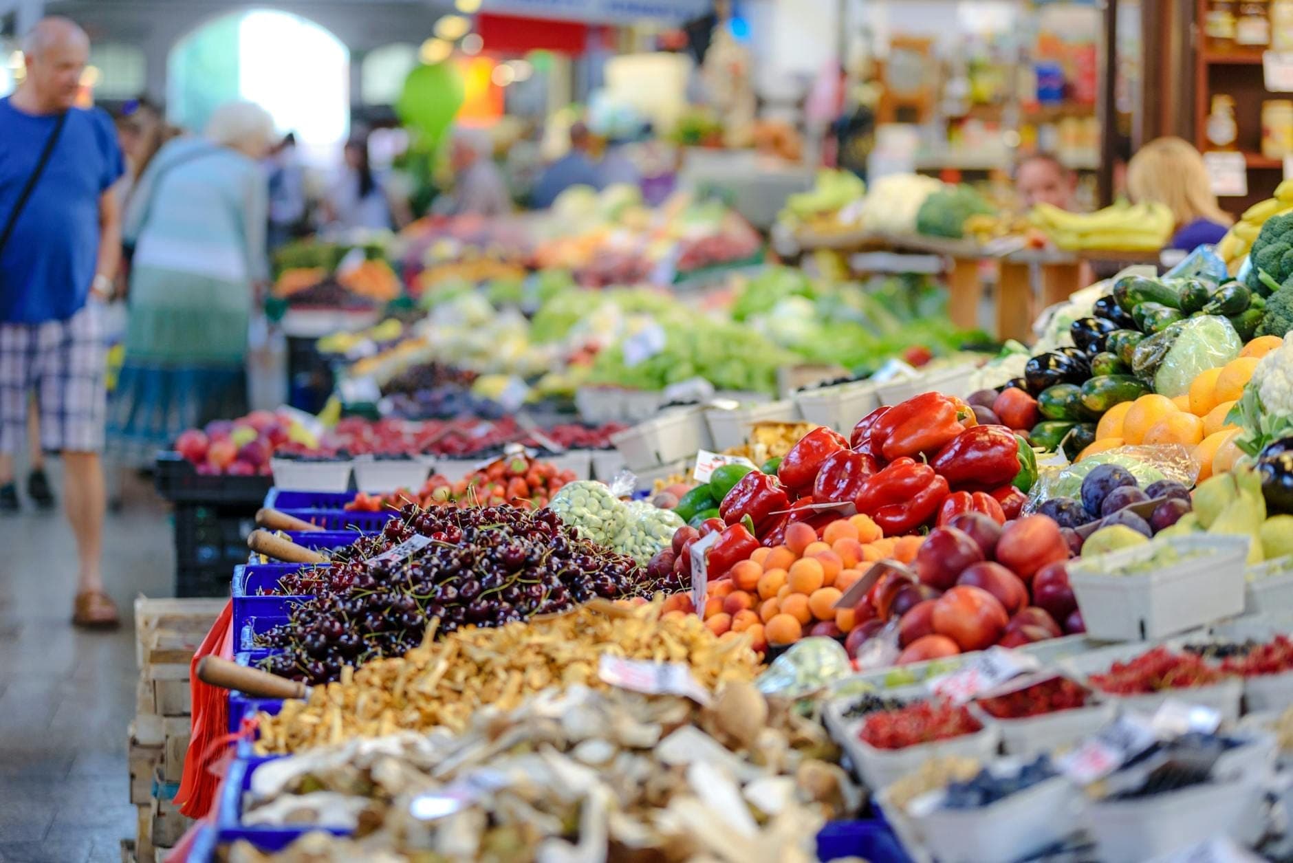 Vivace mercato al coperto con bancarelle piene di frutta e verdura fresca, acquirenti in giro e un'atmosfera animata e colorata.