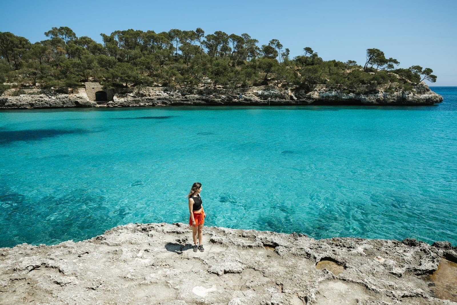 Donna in piedi su una costa rocciosa che si affaccia sulle acque turchesi cristalline di Cala Mondragó a Maiorca, con scogliere coperte di pini sullo sfondo.