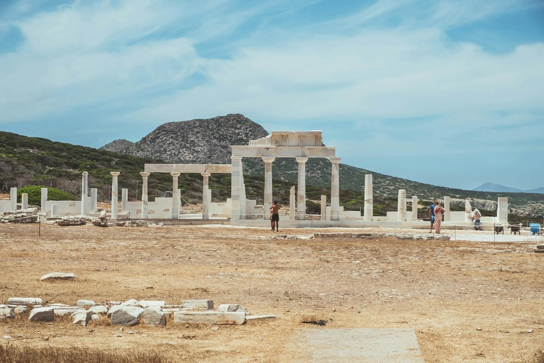 Antike Ruinen mit Säulen und Touristen auf der archäologischen Stätte Delos unter einem blauen Himmel mit Bergen im Hintergrund.