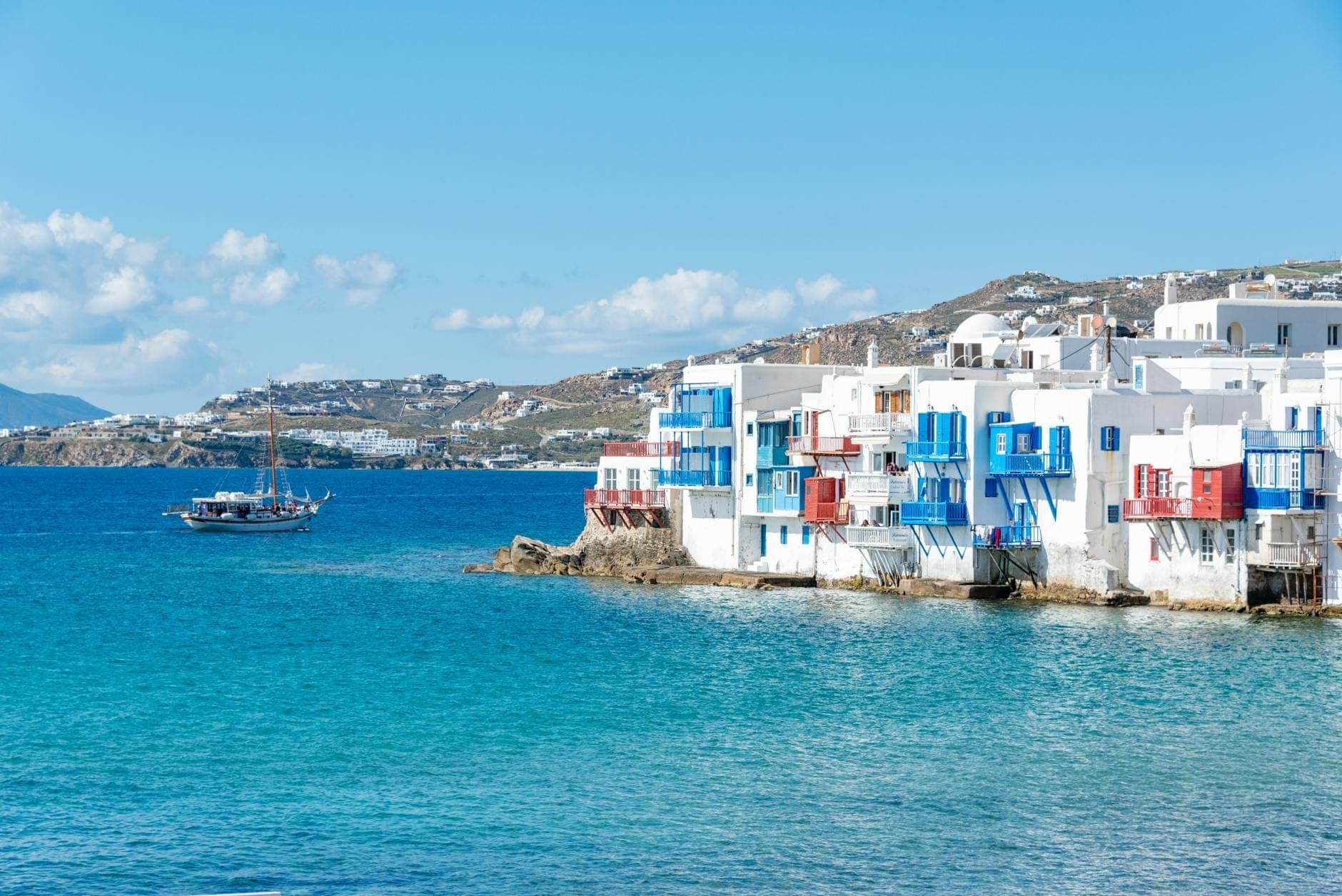 Tiefblaues Meer mit ikonischen weißen Kykladenhäusern und bunten Balkonen an der Uferpromenade von Mykonos, klarer Himmel und sonnenbeschienene Hügel in der Ferne.