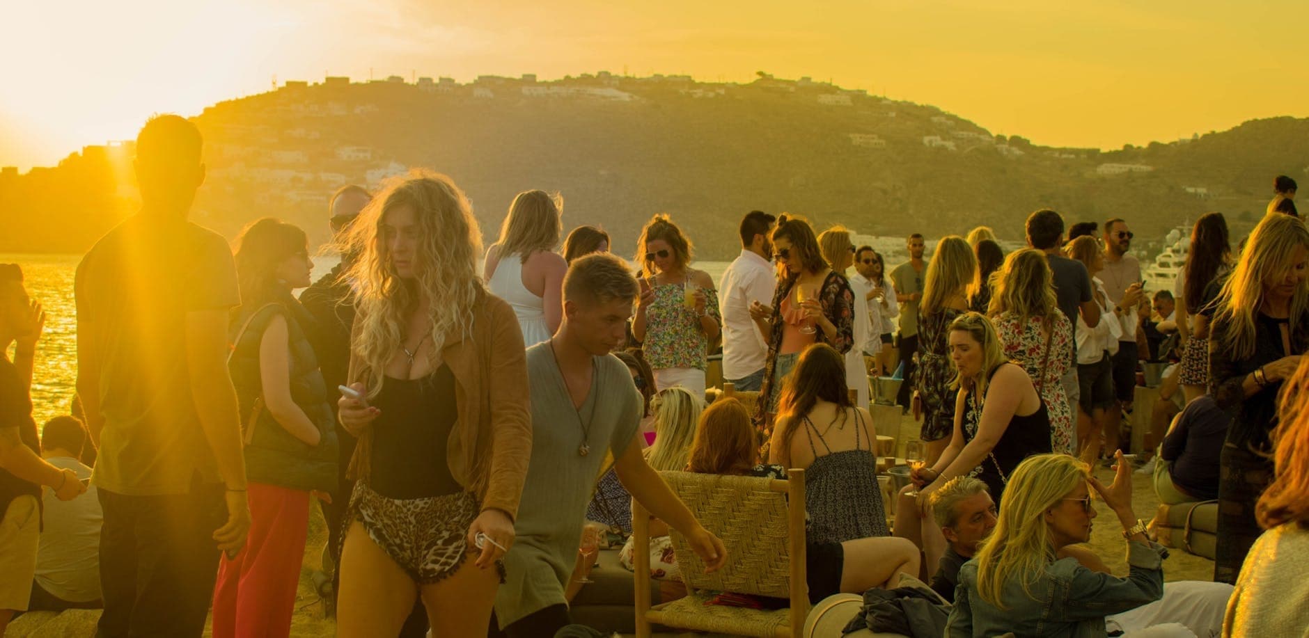 Eine lebhafte Menge genießt Getränke und Musik in einem belebten Beach Club in Mykonos, während die Sonne hinter den Hügeln im Hintergrund untergeht.