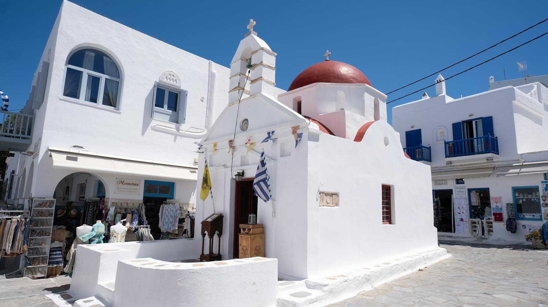 Kleine weiß getünchte Kirche mit roter Kuppel, griechischen Flaggen und nahe gelegenen Geschäften auf einer ruhigen Straße in Mykonos unter klarem blauem Himmel.