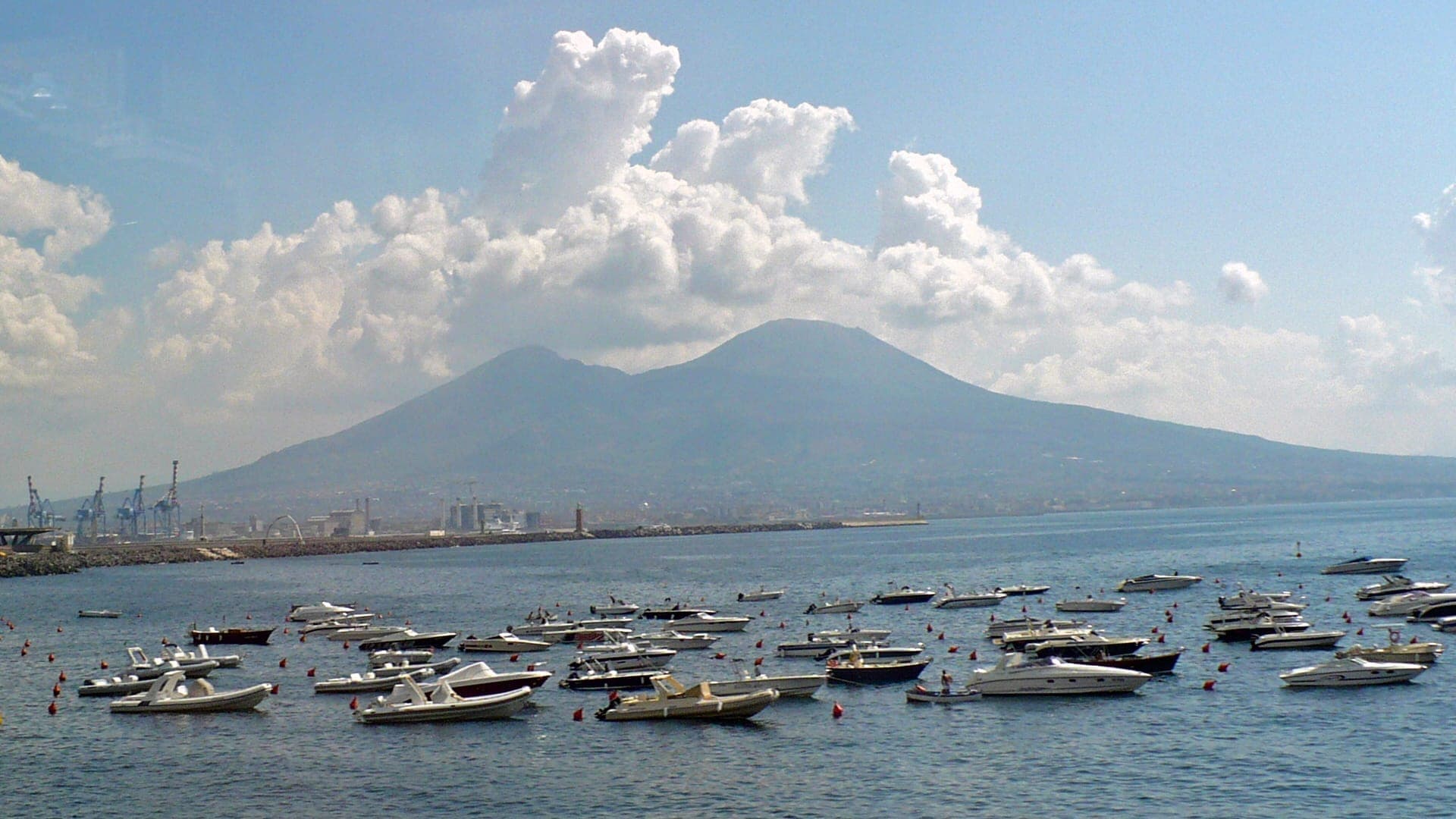 Vista da Baía de Nápoles com barcos na água e o monte Vesúvio ao fundo sob um céu nublado.