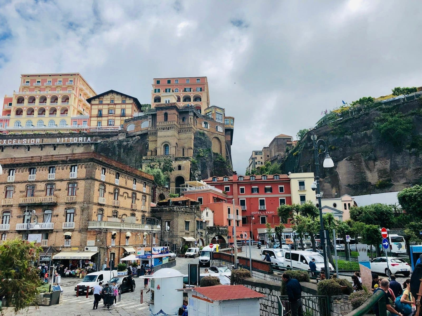 Edifícios históricos coloridos e hotéis, incluindo o Hotel Excelsior Vittoria, situados contra penhascos rochosos no centro de Nápoles sob um céu nublado.