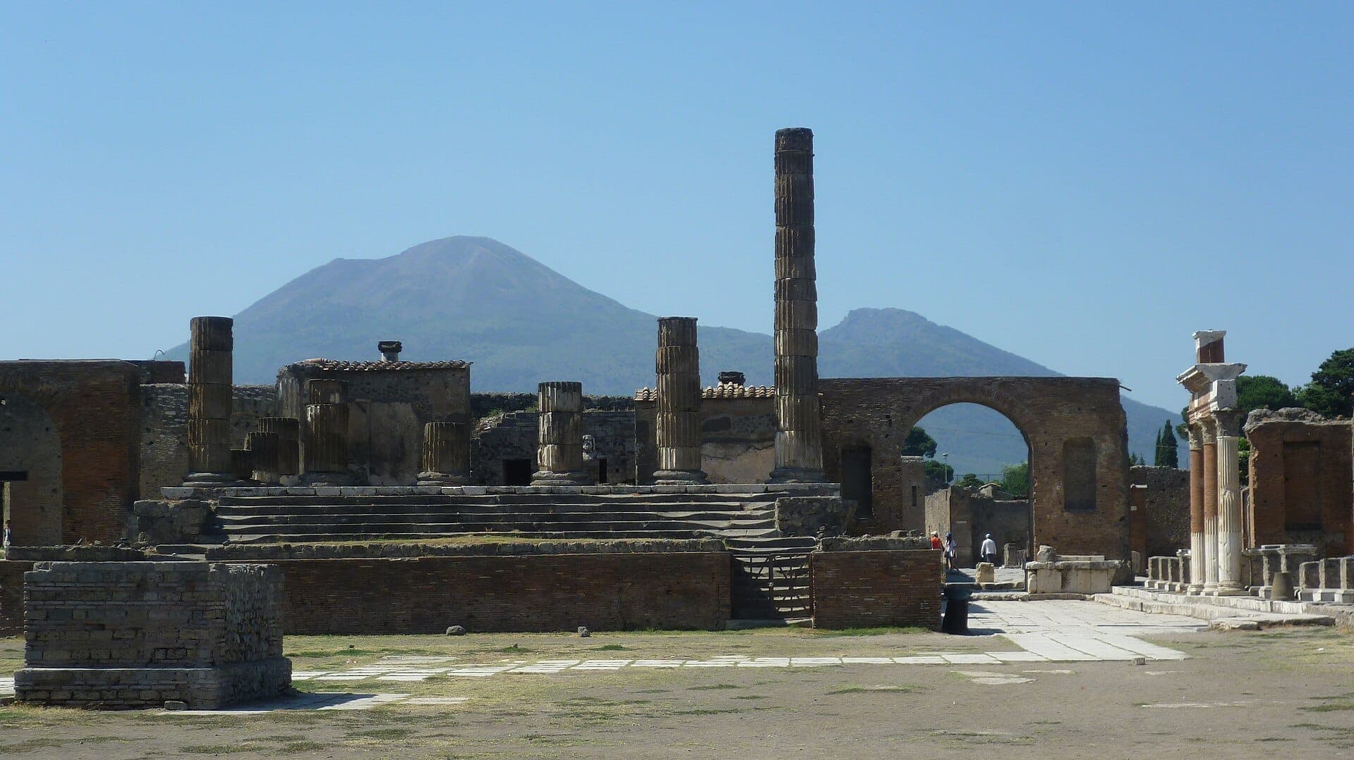 Ruínas de Pompeia com colunas antigas e estruturas de pedra, o Monte Vesúvio ao fundo sob um céu azul claro.