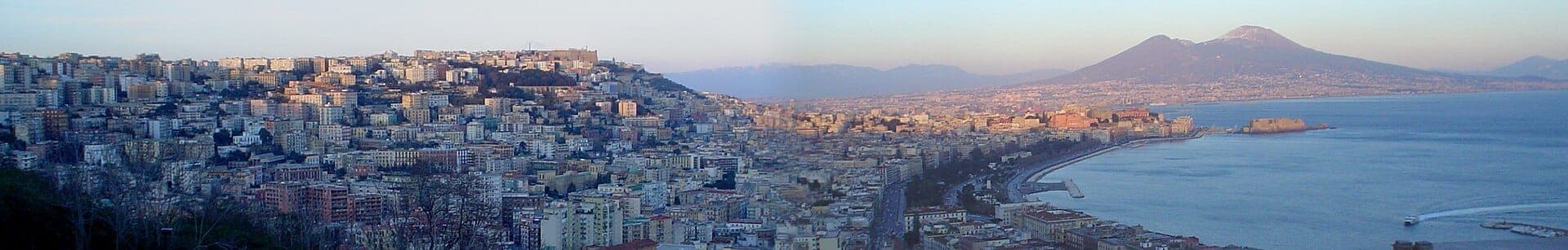 Ampla vista panorâmica sobre Nápoles com edifícios da cidade, a orla marítima, a baía e o Monte Vesúvio ao fundo sob um céu limpo.