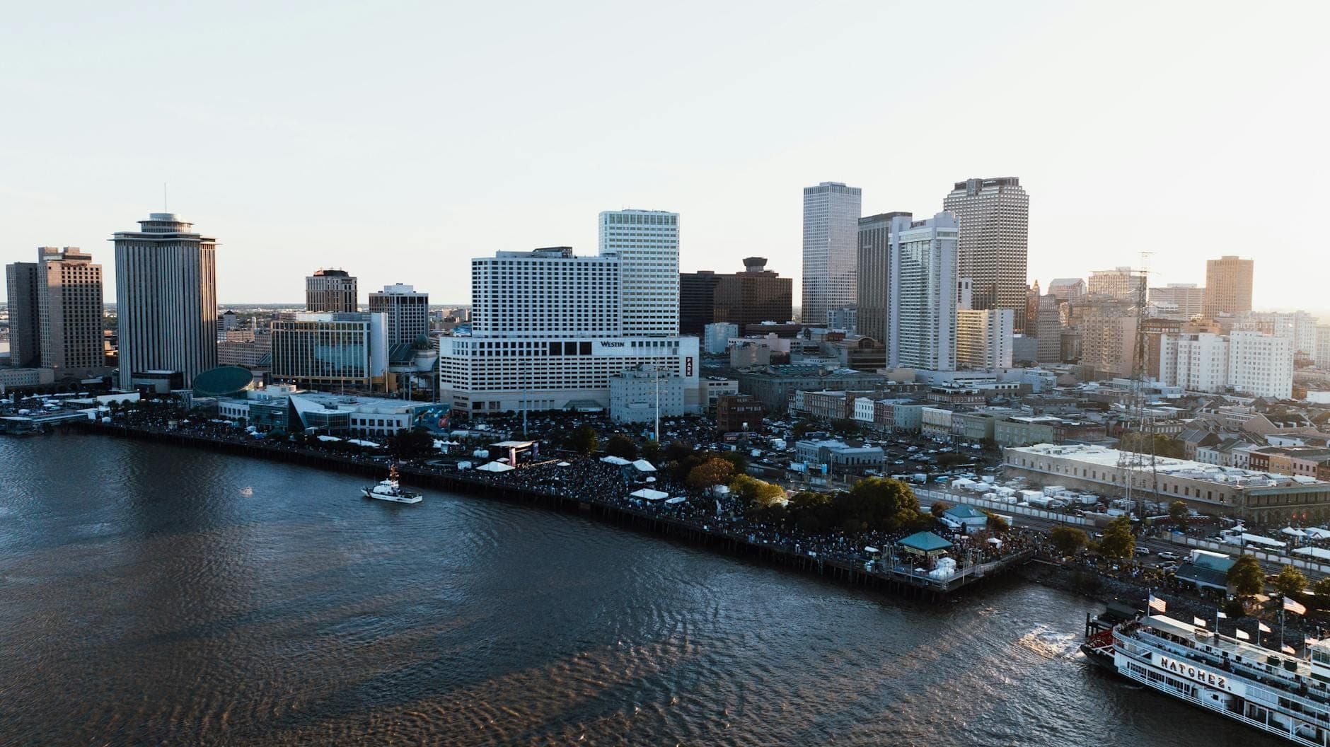Vista aérea do orla fluvial de New Orleans com o horizonte da cidade, barcos no rio Mississippi e pessoas ao longo do calçadão.