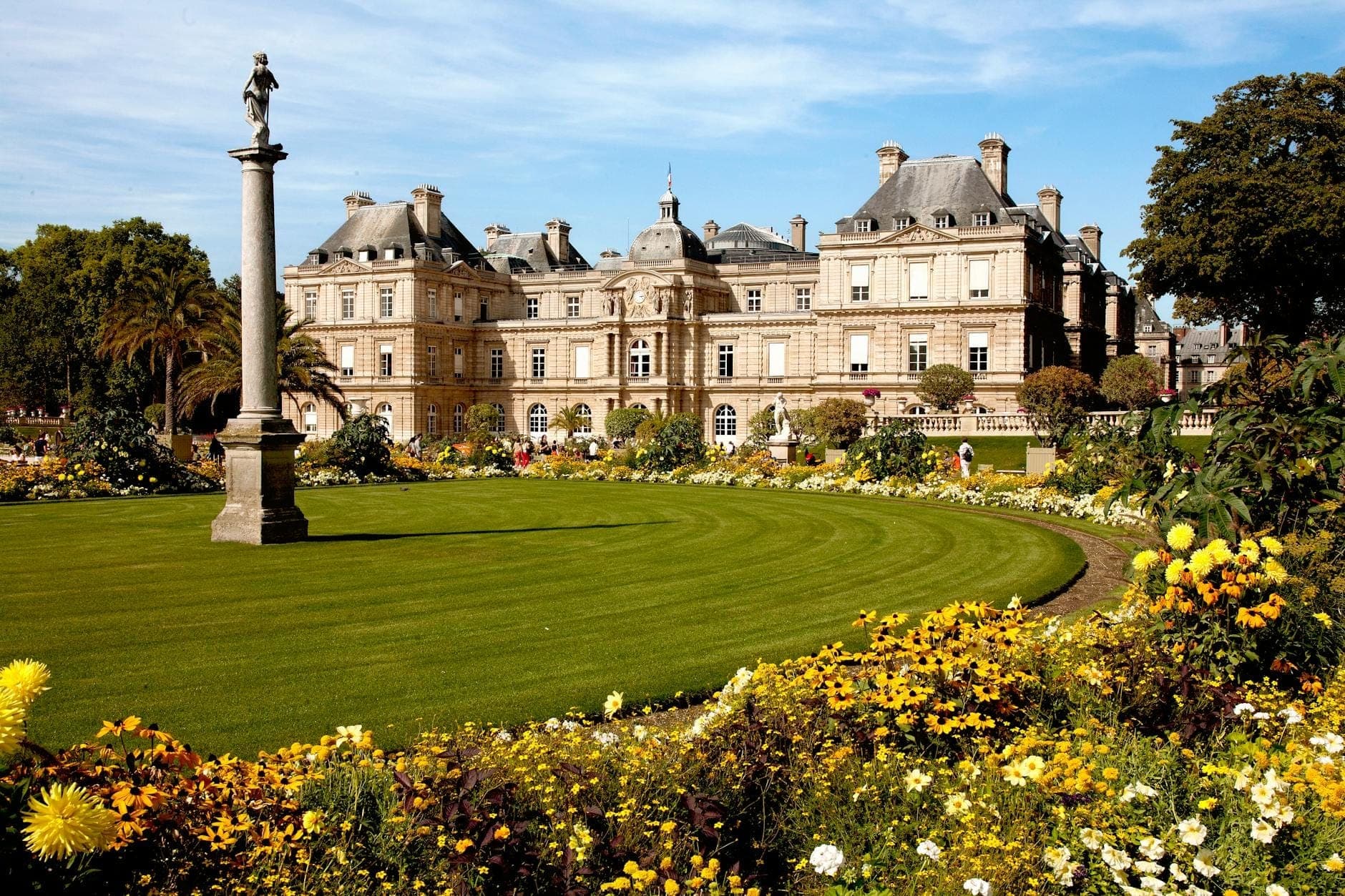 Weitläufiger Blick auf den Jardin du Luxembourg in Paris mit üppig grünem Rasen, bunten Blumen, Statuen und dem prachtvollen Palais du Luxembourg im Hintergrund.