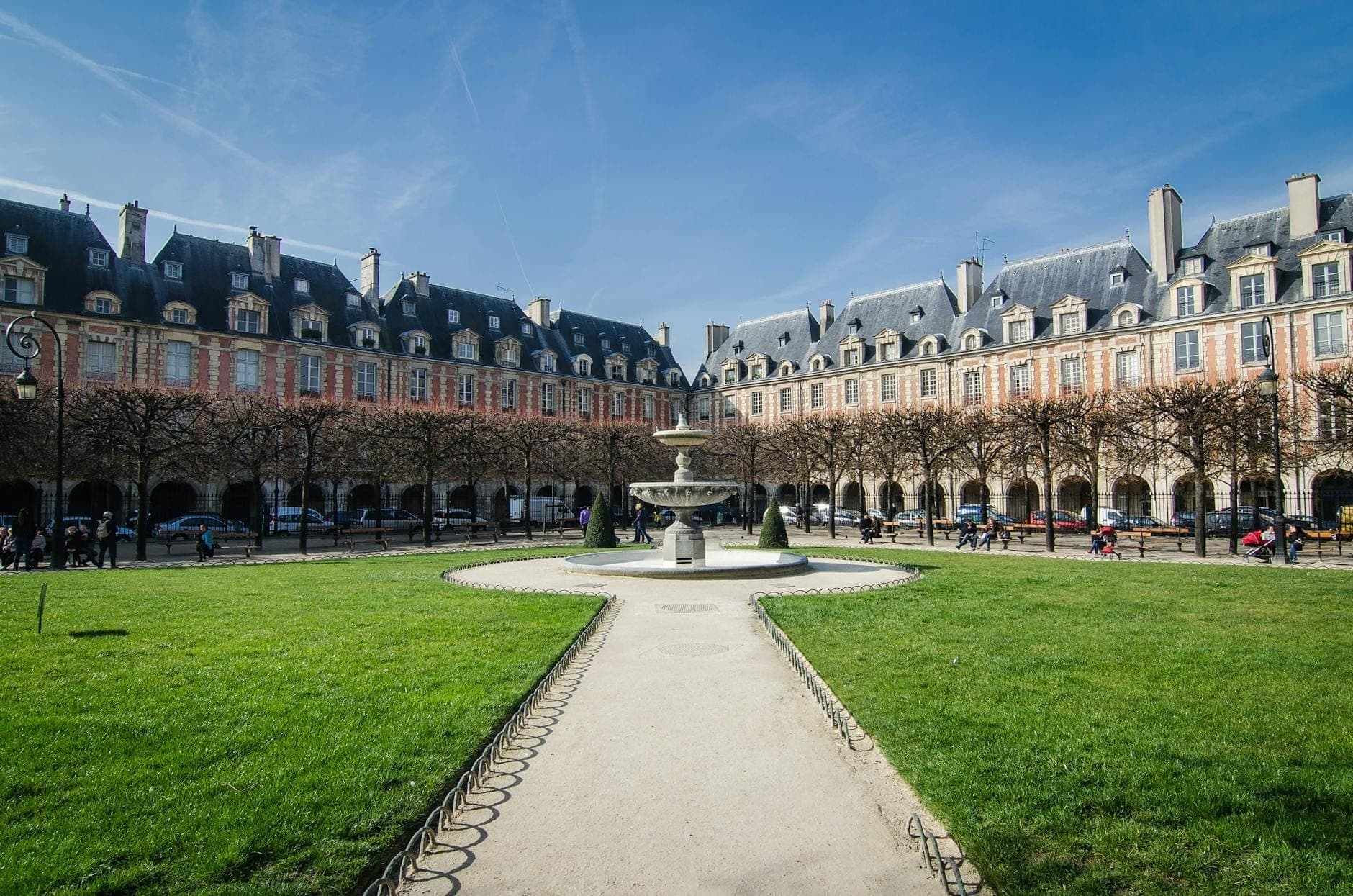 Weitläufige Ansicht der Place des Vosges in Paris mit klassischen Bogenganggebäuden, zentralem Brunnen, grünen Rasenflächen und Menschen, die unter blauem Himmel auf Wegen spazieren.