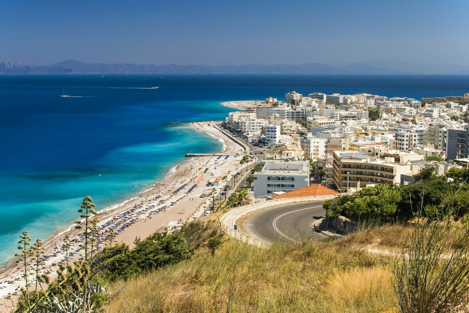 Vue panoramique de la ville de Rhodes avec ses maisons blanches le long d'un littoral incurvé, la mer Égée turquoise et des montagnes au loin sous un ciel bleu limpide.