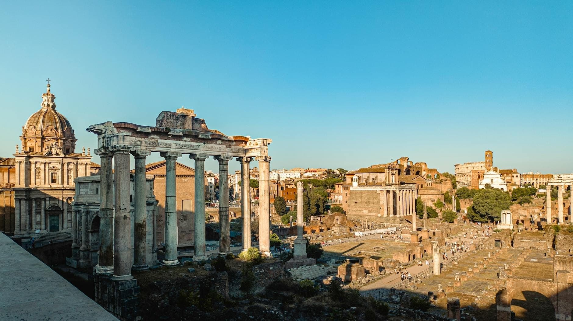 Panoramablick auf das Forum Romanum mit antiken Ruinen, Säulen und umliegenden historischen Gebäuden unter einem klaren blauen Himmel.