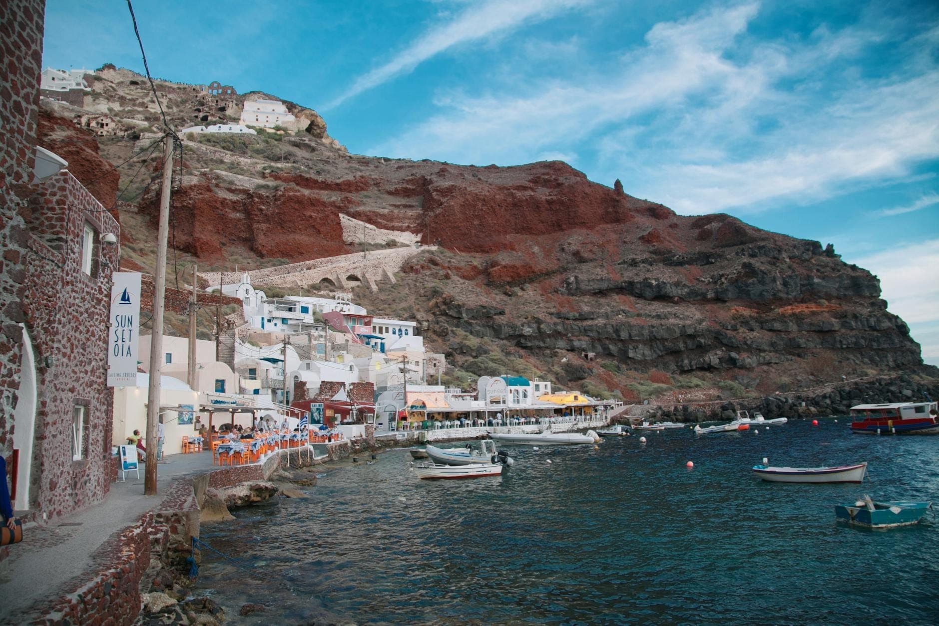 Piccole barche ancorate vicino alla riva di Santorini con scogliere vulcaniche ed edifici imbiancati, che mostrano la caratteristica costa rocciosa.