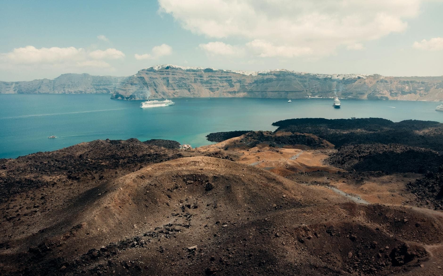 Paesaggio vulcanico di Santorini con vista sulla caldera, barche e nave da crociera sull'acqua blu sotto un cielo parzialmente nuvoloso.