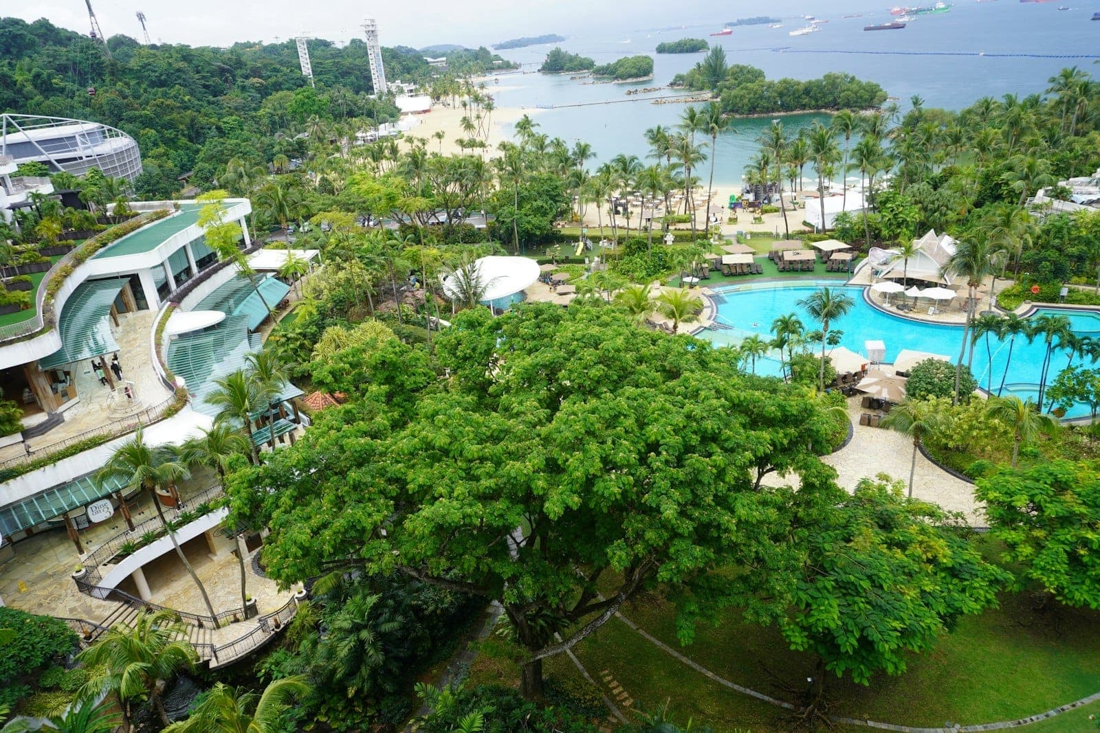 มุมมองทางอากาศของ Sentosa Island ที่แสดงให้เห็นพืชพรรณเขียวขจี สระว่ายน้ำของรีสอร์ท หาดทราย และทะเลพร้อมเรือที่จอดทอดสมออยู่เบื้องหลัง