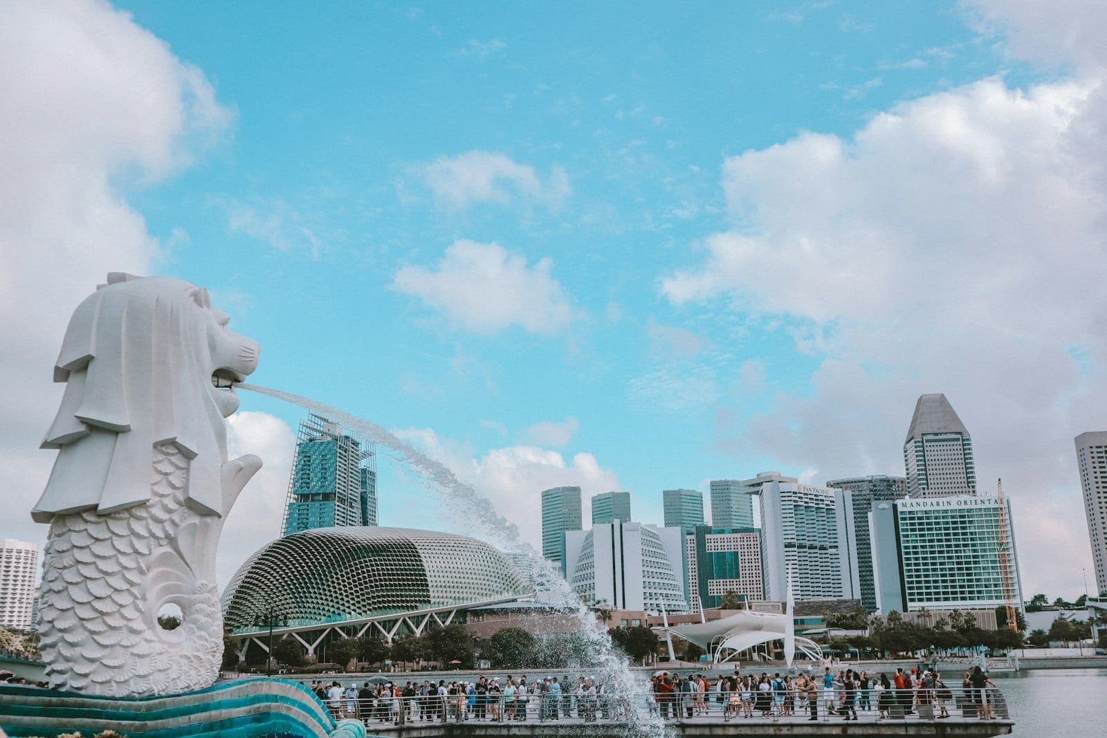 รูปปั้นเมอร์ไลออนพ่นน้ำอันเป็นสัญลักษณ์ โดยมีฉากหลังเป็นสกายไลน์สมัยใหม่และแลนด์มาร์คชื่อดังของสิงคโปร์ภายใต้ท้องฟ้าสดใสมีเมฆบางส่วน