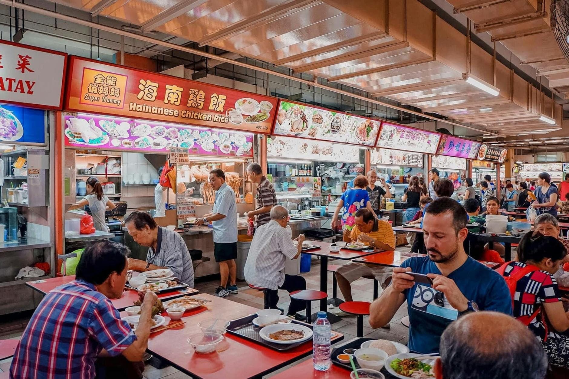 ศูนย์อาหาร Hawker Centre ที่賑คึกคักในสิงคโปร์ มีนักทานนั่งรับประทานอาหารที่โต๊ะและแผงอาหารสีสันสดใสที่ขายเมนูอย่างข้าวไก่ไหหลำ
