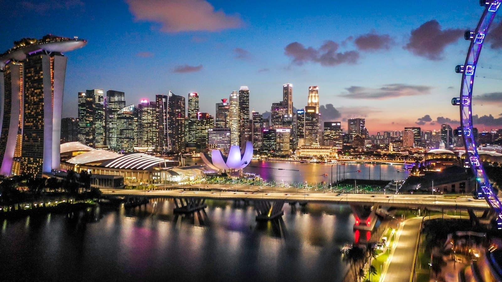 วิวยามพลบค่ำอันสดใสของ Marina Bay ในสิงคโปร์ โดยมีเส้นขอบฟ้าของเมือง, Marina Bay Sands, Helix Bridge, ArtScience Museum และ Singapore Flyer ที่มองเห็นได้ชัดเจน
