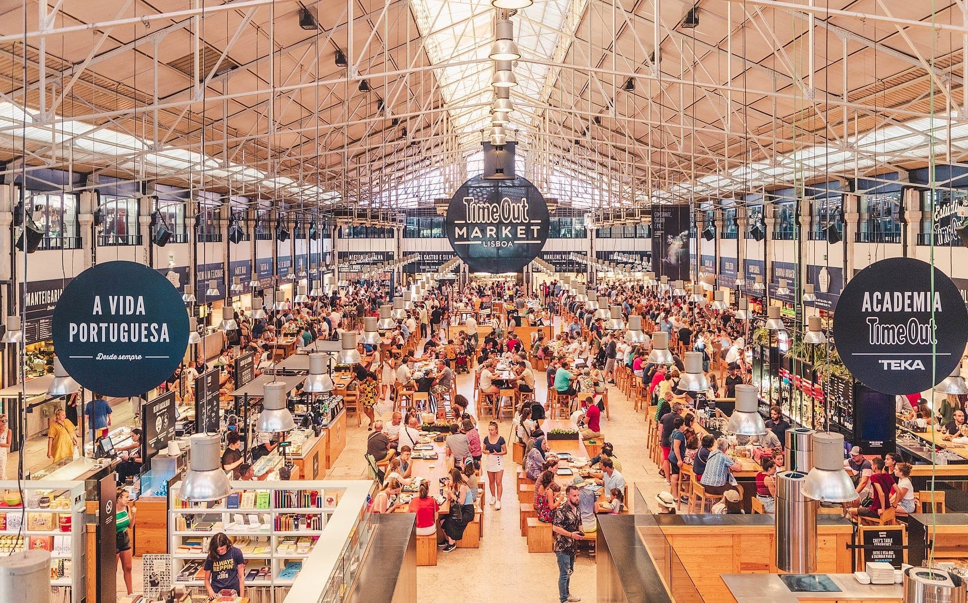 Vista interior ampla do Time Out Market Lisboa, mostrando multidões movimentadas, mesas coletivas de madeira, bancas de comida e teto de vidro com estrutura de ferro.
