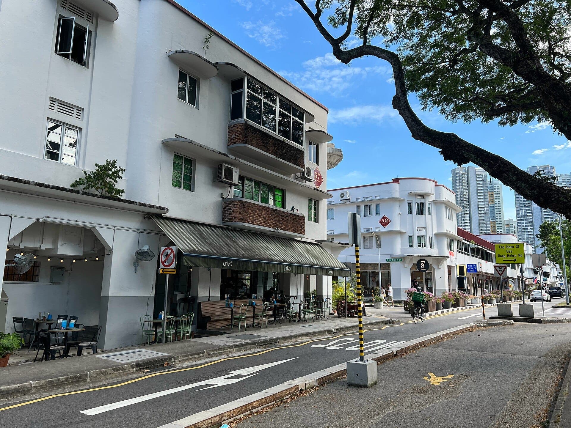 อาคารสีขาวสไตล์ Streamline Moderne และร้านกาแฟเรียงรายตามถนนเงียบสงบใน Tiong Bahru ท่ามกลางต้นไม้เขียวชอุ่มและท้องฟ้าสีฟ้า