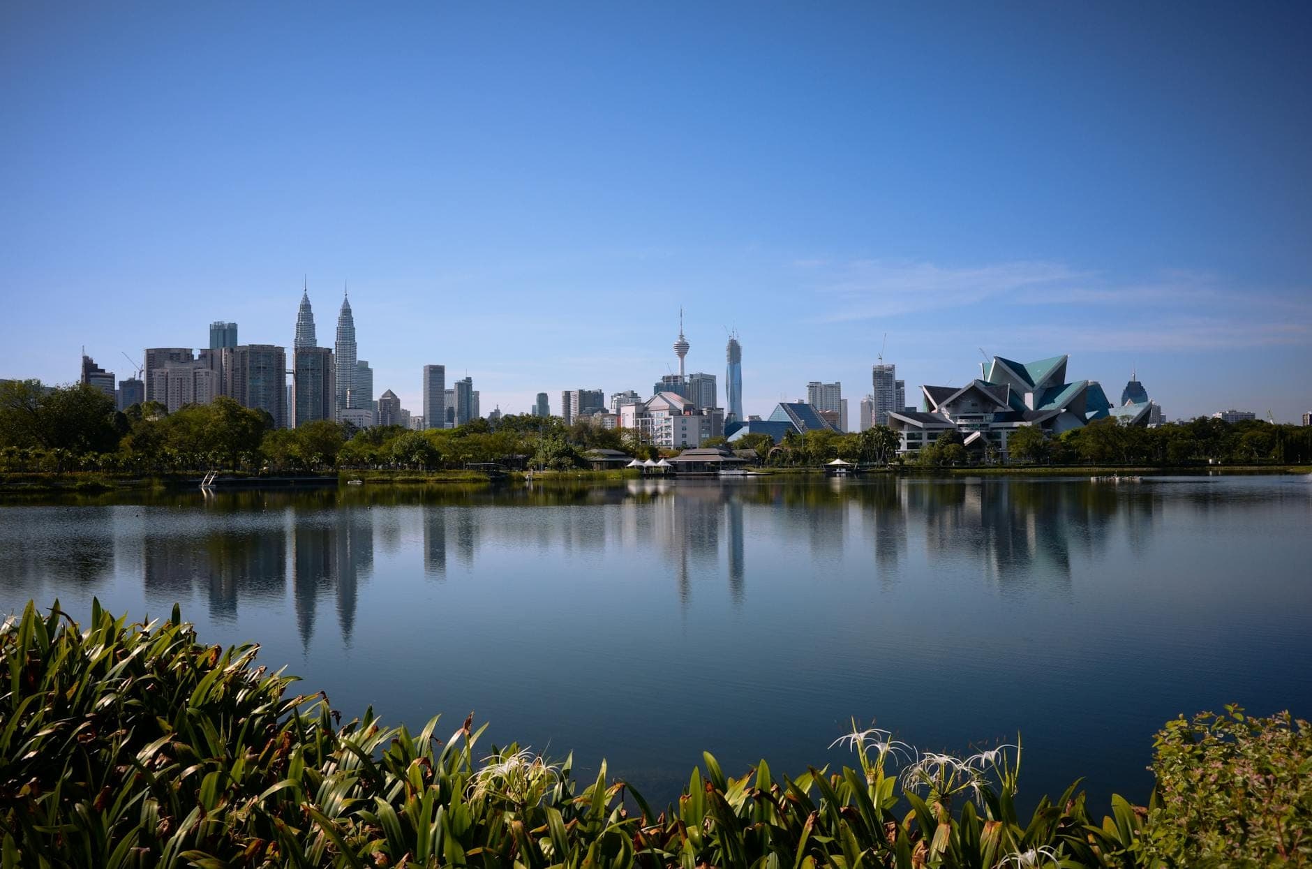 Lac du Parc Titiwangsa avec les reflets de la skyline de Kuala Lumpur, incluant les Tours Petronas et l'architecture moderne, sous un ciel bleu dégagé.