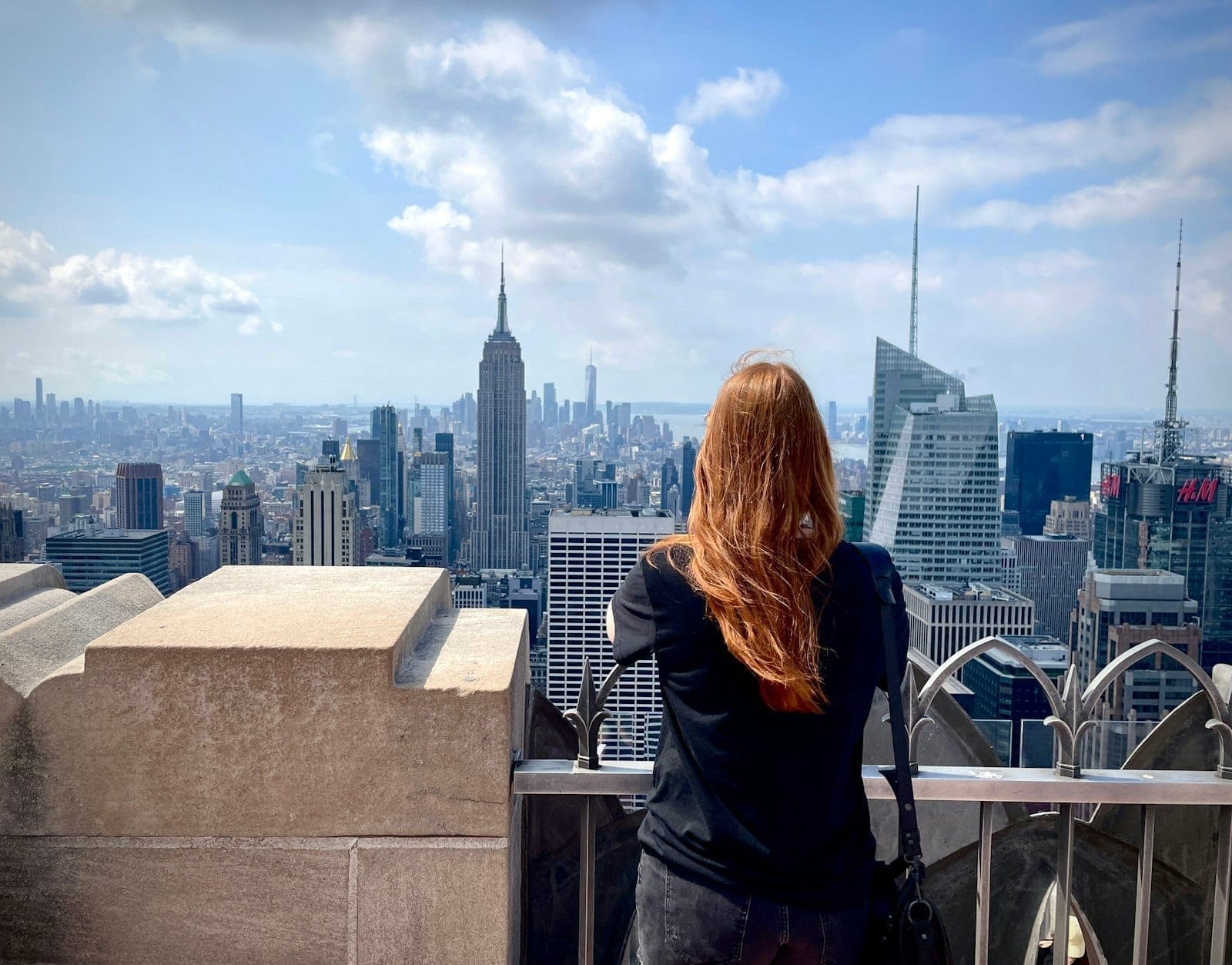 หญิงผมแดงยืนชมวิวเส้นขอบฟ้าแมนฮัตตันจาก Top of the Rock เห็นตึก Empire State ชัดเจนในวันฟ้าครึ้มบางส่วน