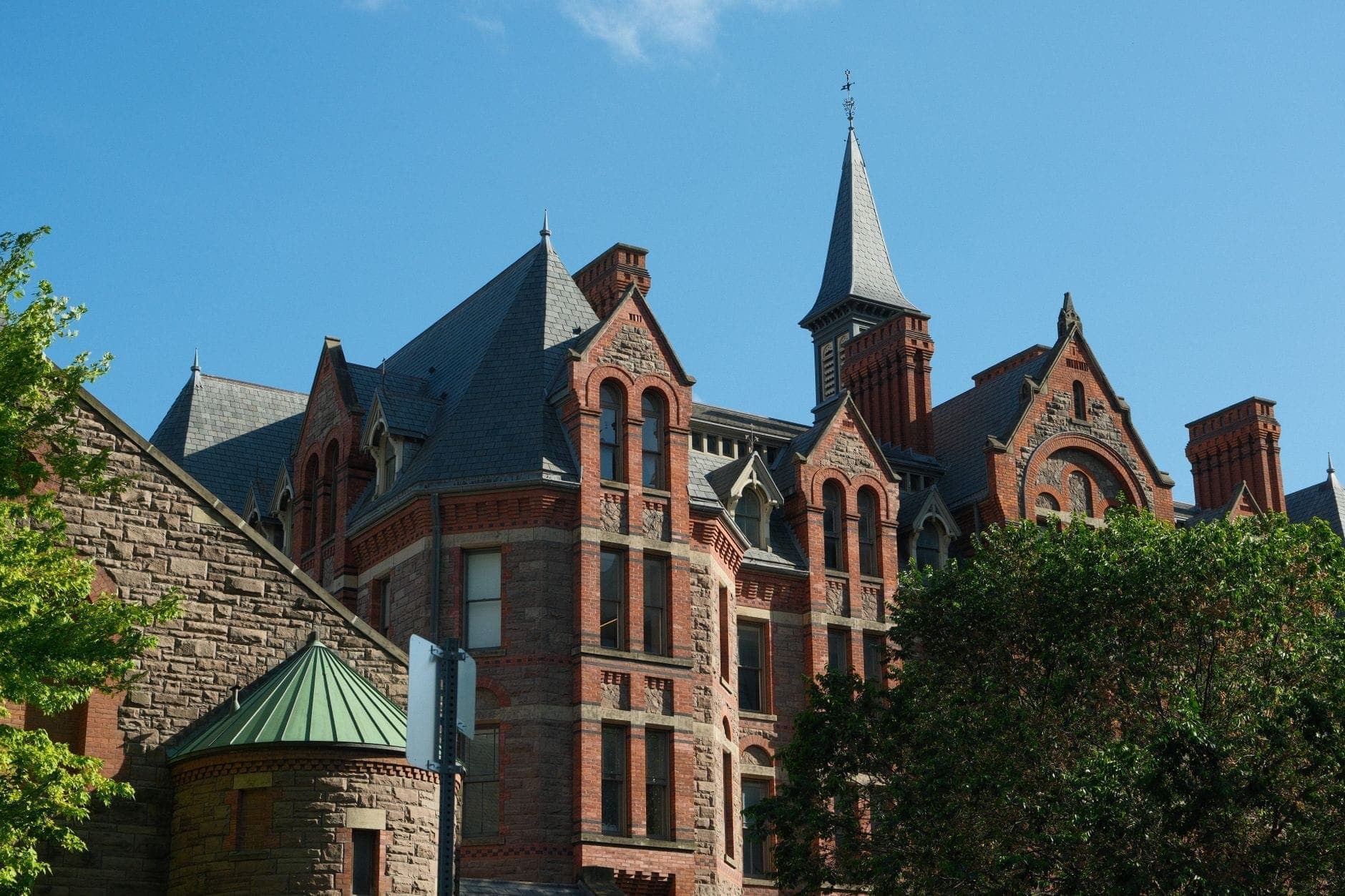 Victorian-era red-brick building with pointed roofs and turrets against a blue sky and trees in Toronto.
