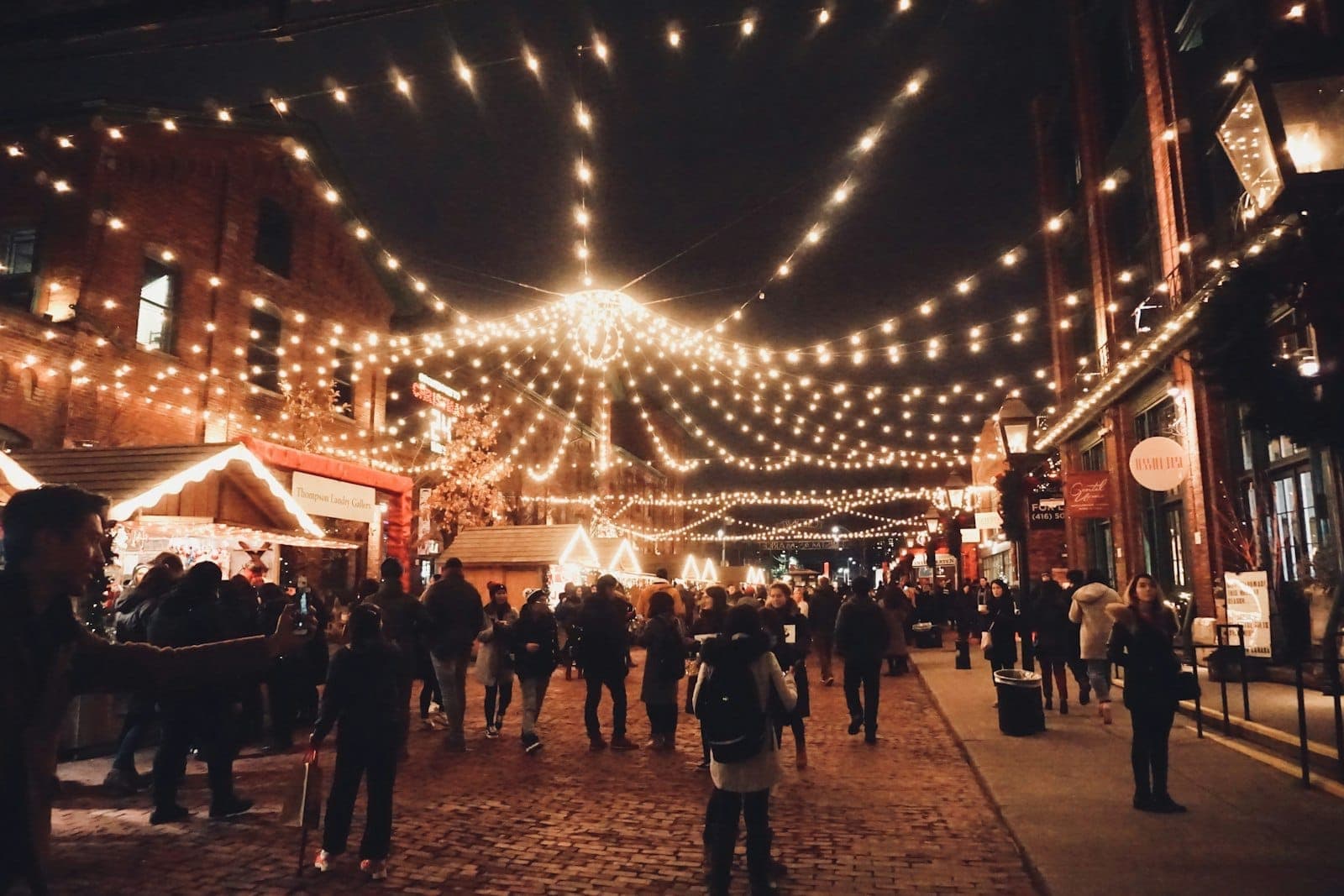 Una folla di persone passeggia tra i vicoli in acciottolato del Distillery District di Toronto di notte, immersa tra luci festive e casette di legno del mercatino.