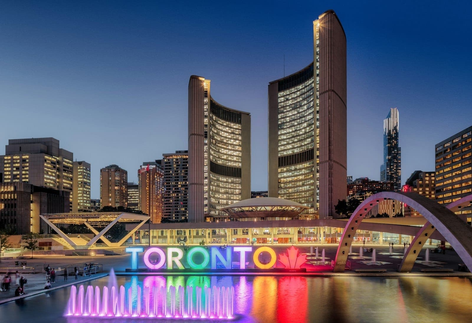 Il Toronto City Hall al crepuscolo con le due torri curve illuminate, la cupola, la colorata insegna Toronto e la fontana di Nathan Phillips Square.